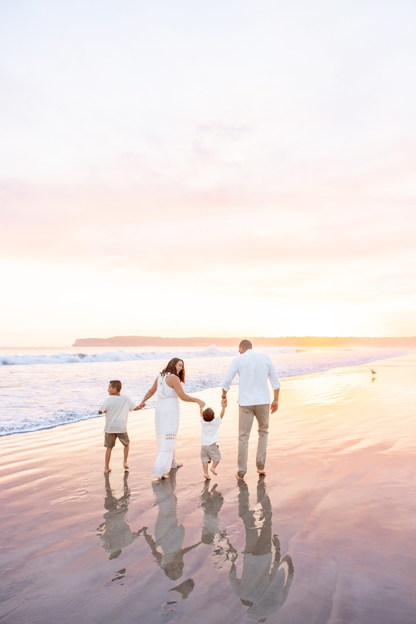 A happy family in white outfits hold hands and walk on a beach at sunset before visiting restaurants with play areas in san diego