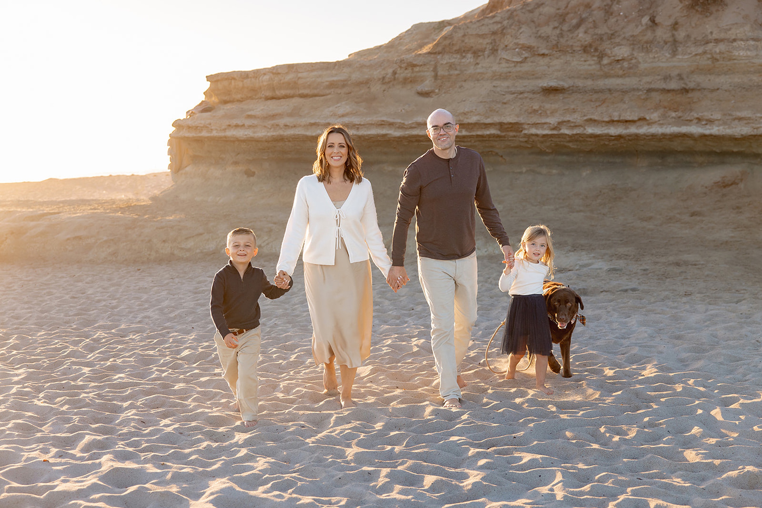 Happy mom and dad walk on a beach at sunset with their toddler son and daughter and brown dog