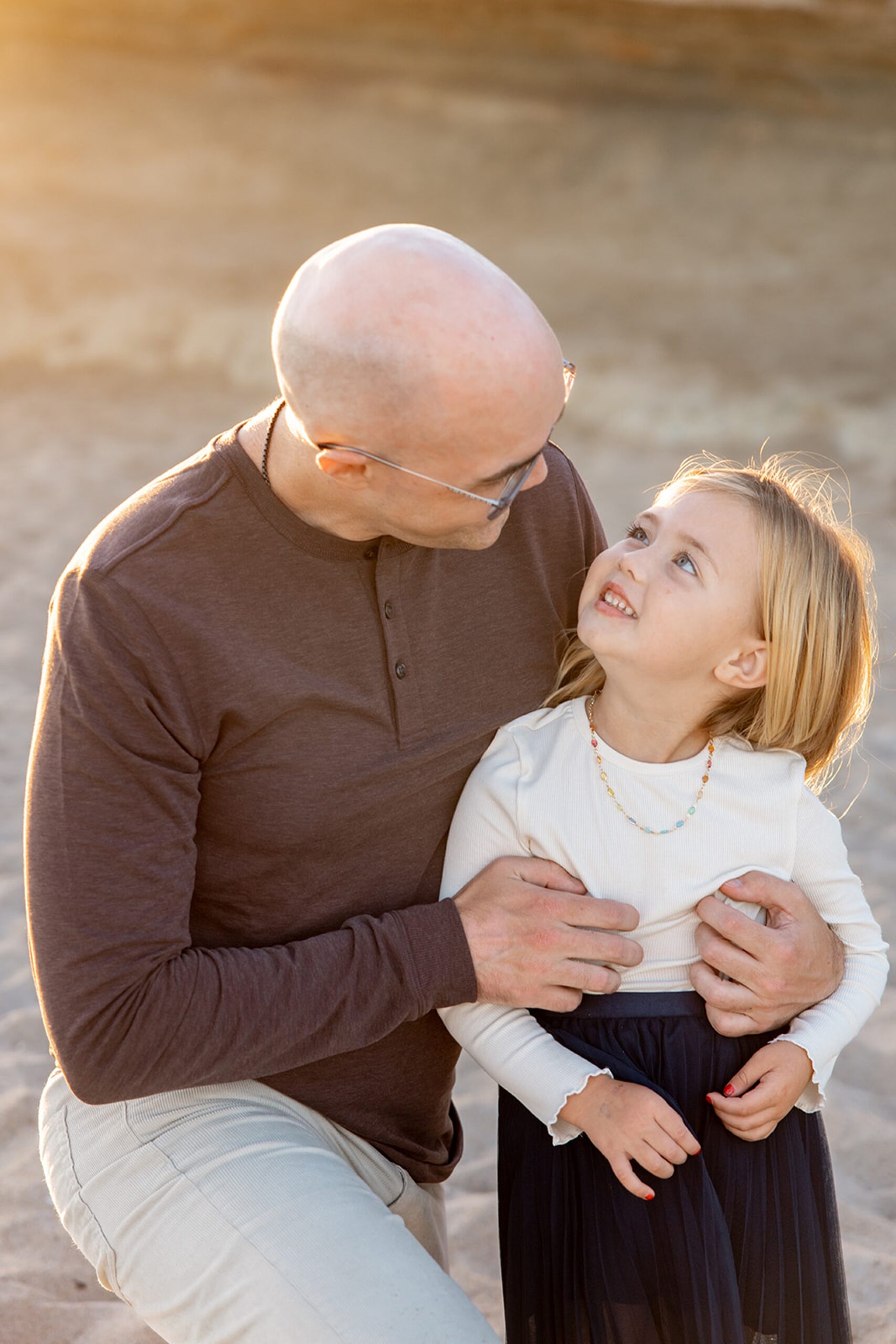 A dad in a brown shirt hugs and smiles at his toddler daughter on a beach in a white and black dress during things to do in San Diego with toddlers