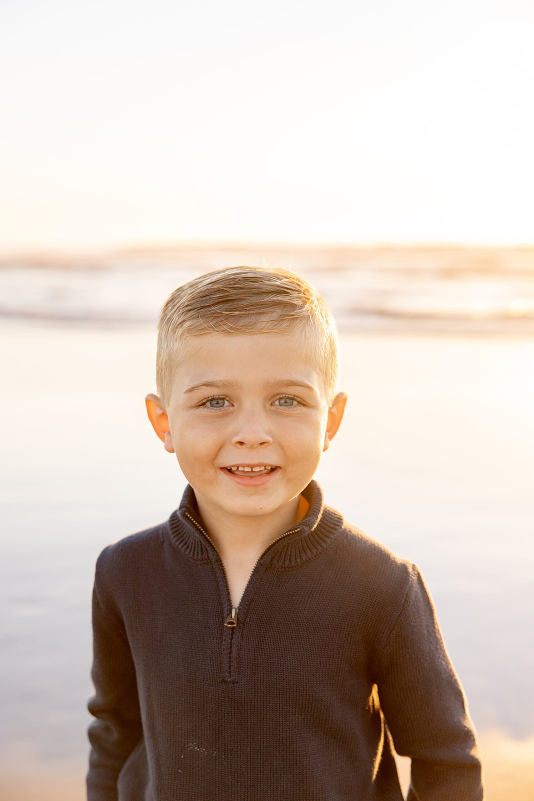 A young boy in a brown sweater smiles on a beach at sunset during things to do in San Diego with toddlers