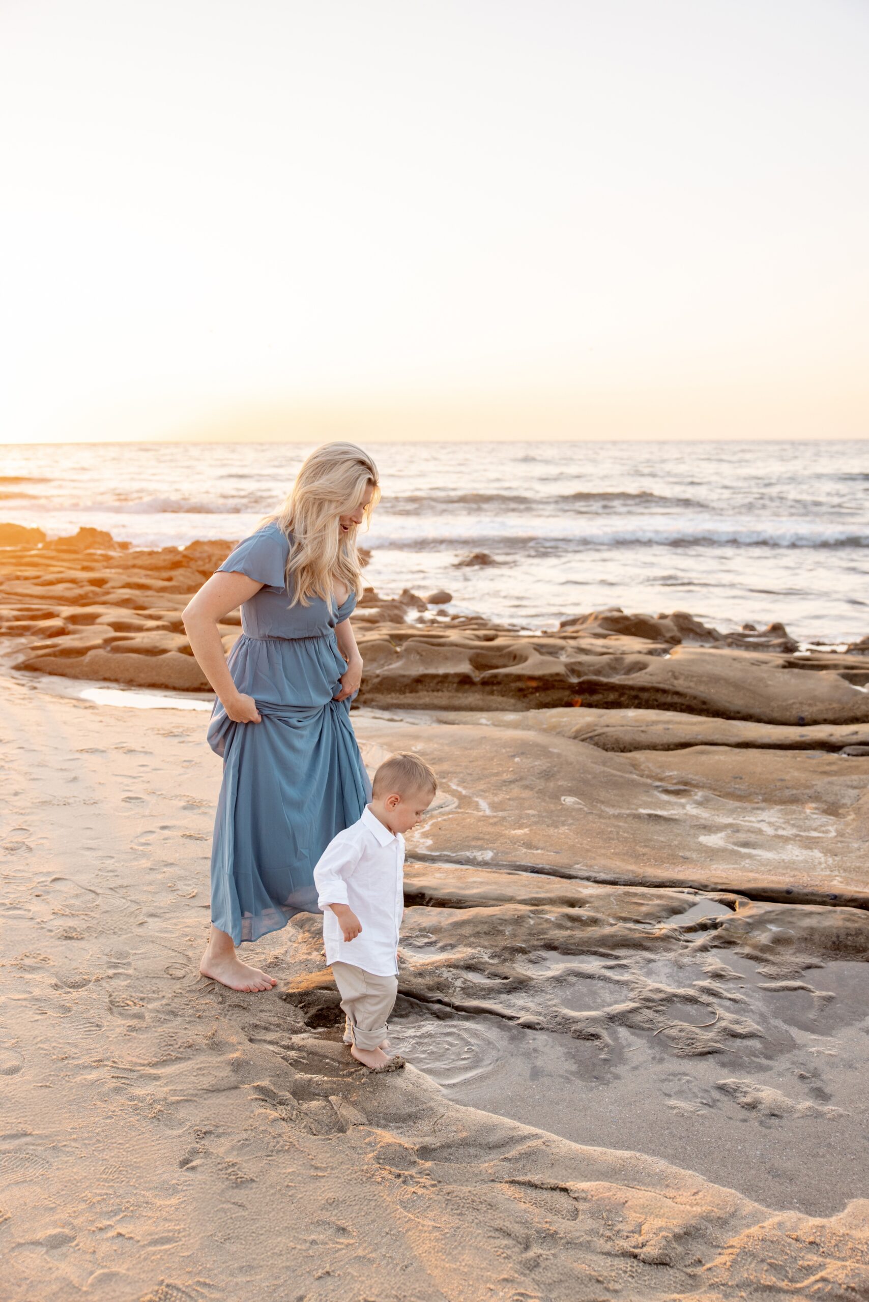 A mom in a blue dress explores a tide pool on the beach with her toddler in a white shirt and khakis