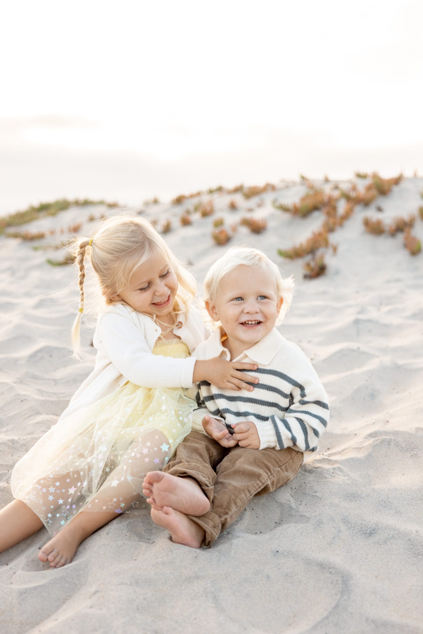 A toddler girl in a yellow dress hugs her baby brother in a stripe sweater while sitting on a beach before visiting toddler-friendly restaurants in San Diego