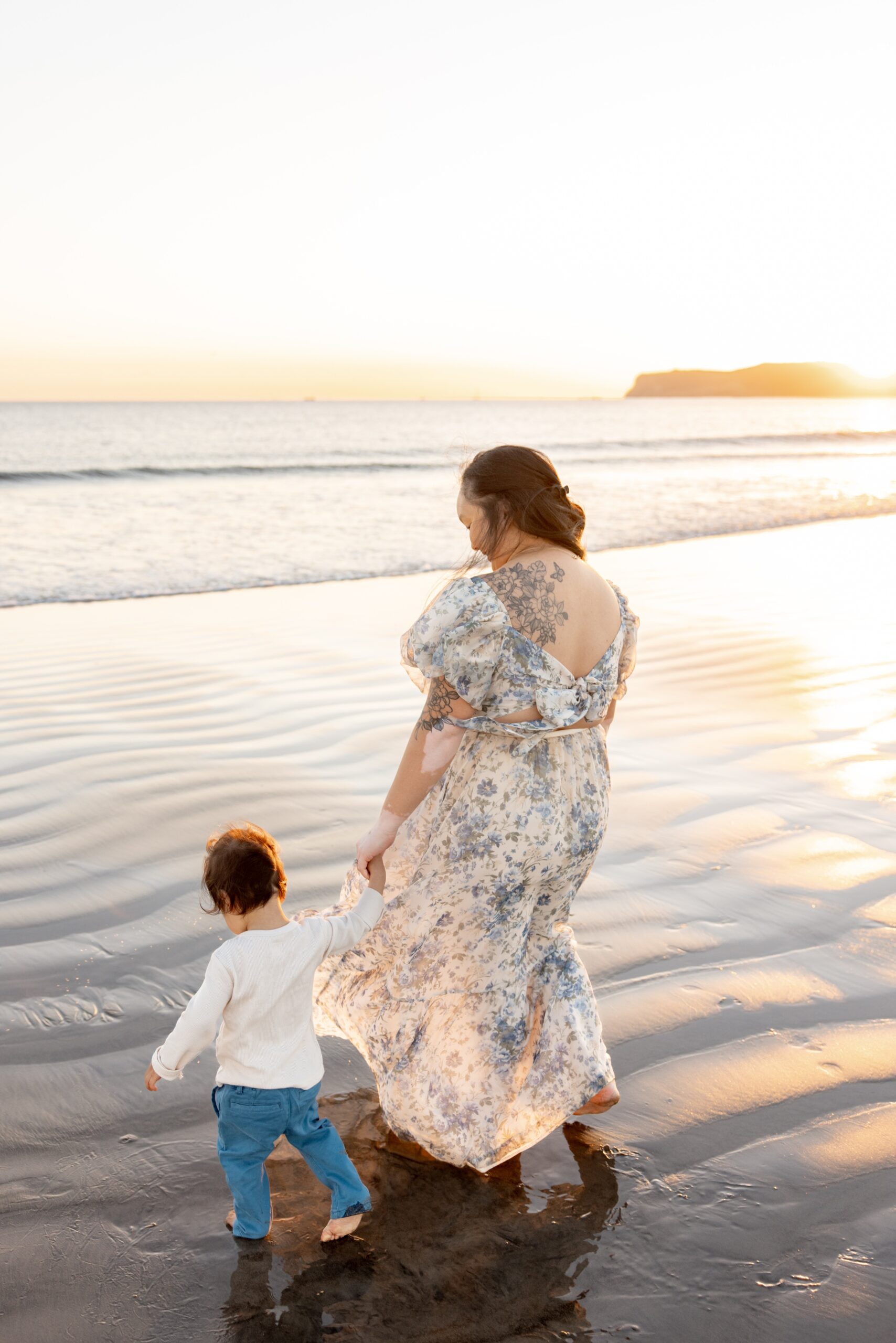 A mother walks her toddler on the beach at sunset holding hands