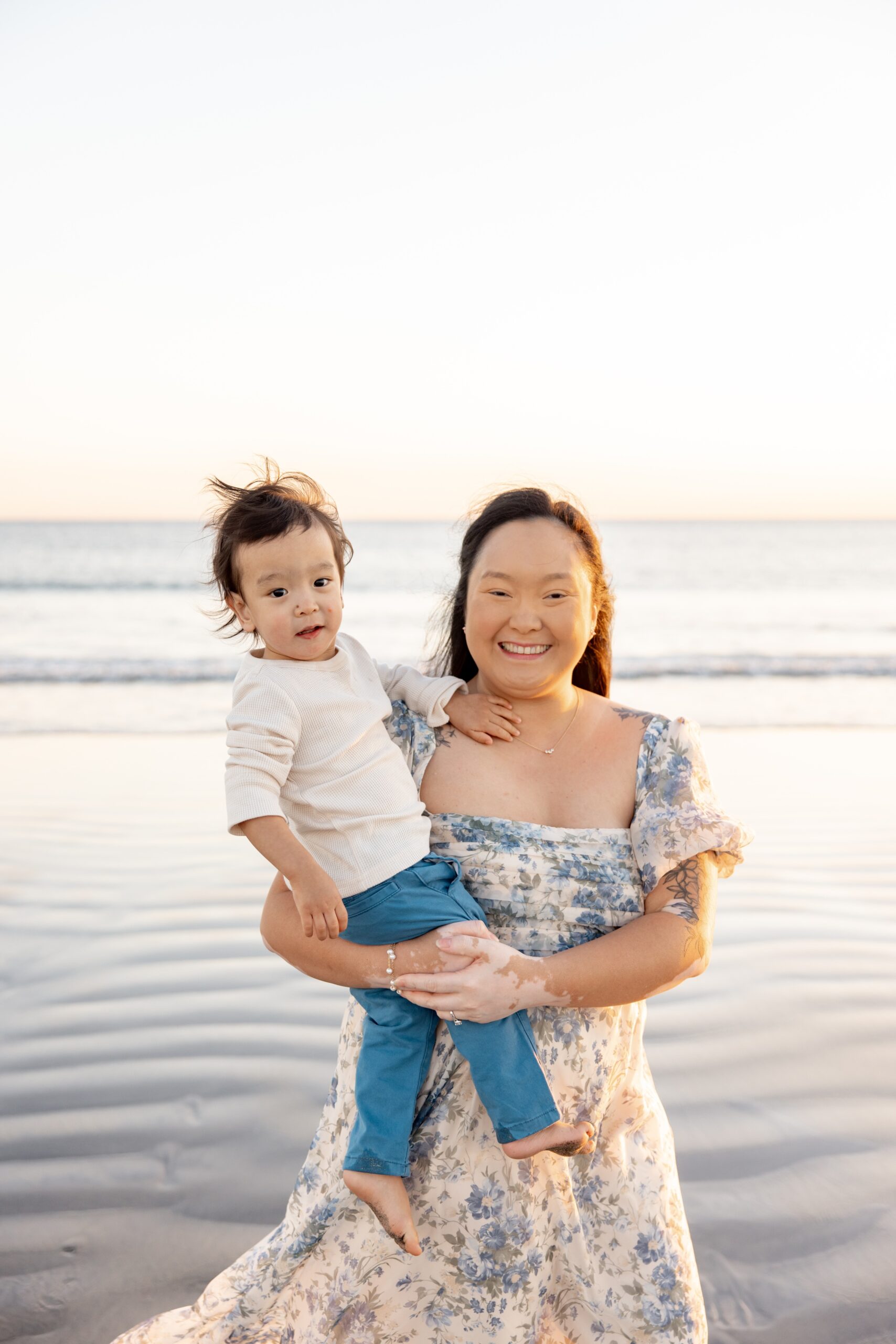 A smiling mother stands on a beach holding her toddler in a flowing floral print gown after some family events in san diego
