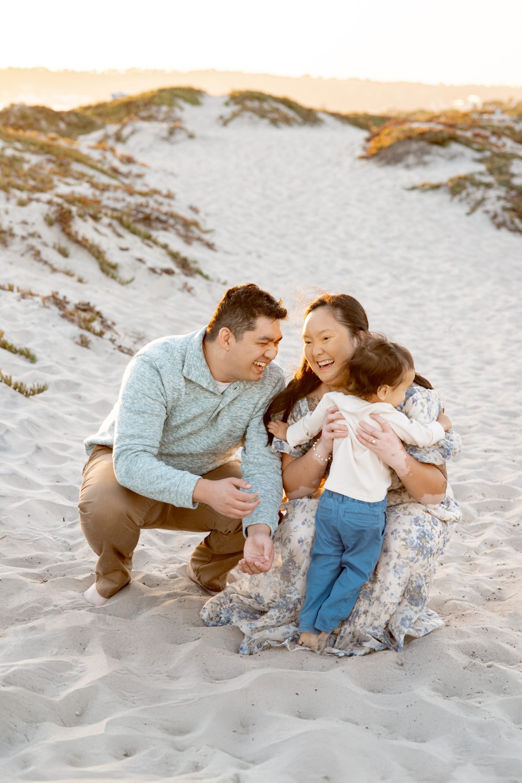 Laughing mom and dad play with their toddler on a beach dune after some family events in san diego