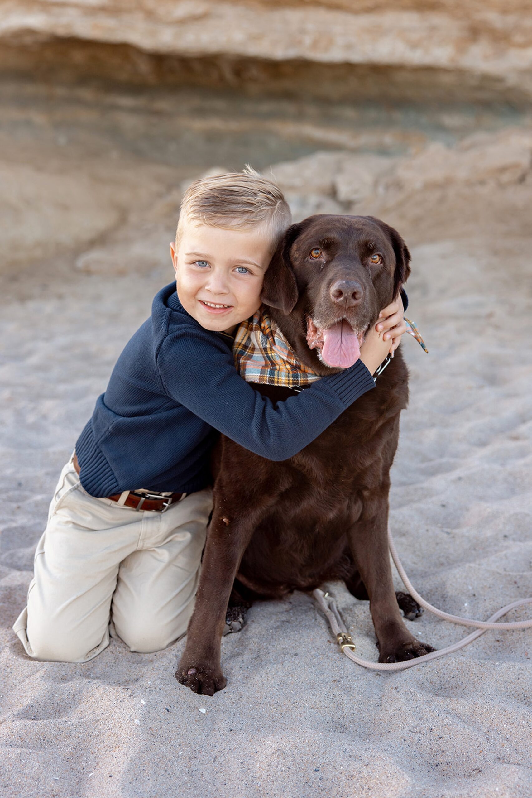A young blonde boy in a blue sweater hugs his chocolate lab on a beach