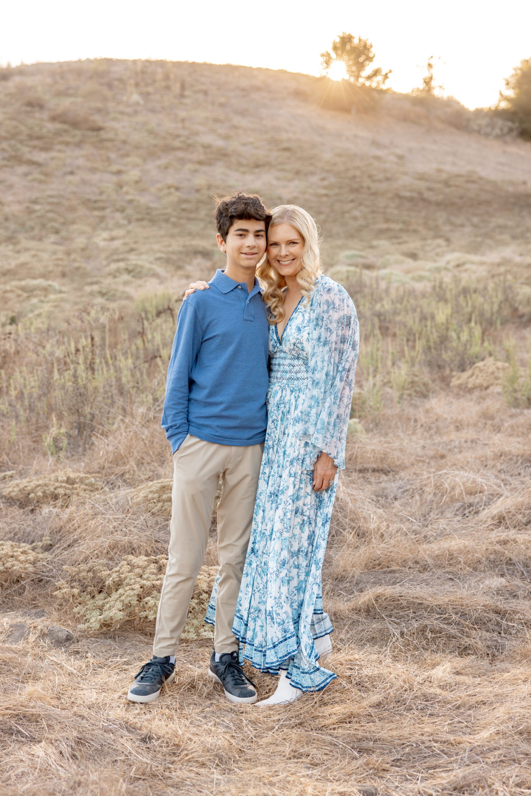 A happy mom in a blue dress stands smiling with her teenage son on a hillside at sunset during family events in san diego
