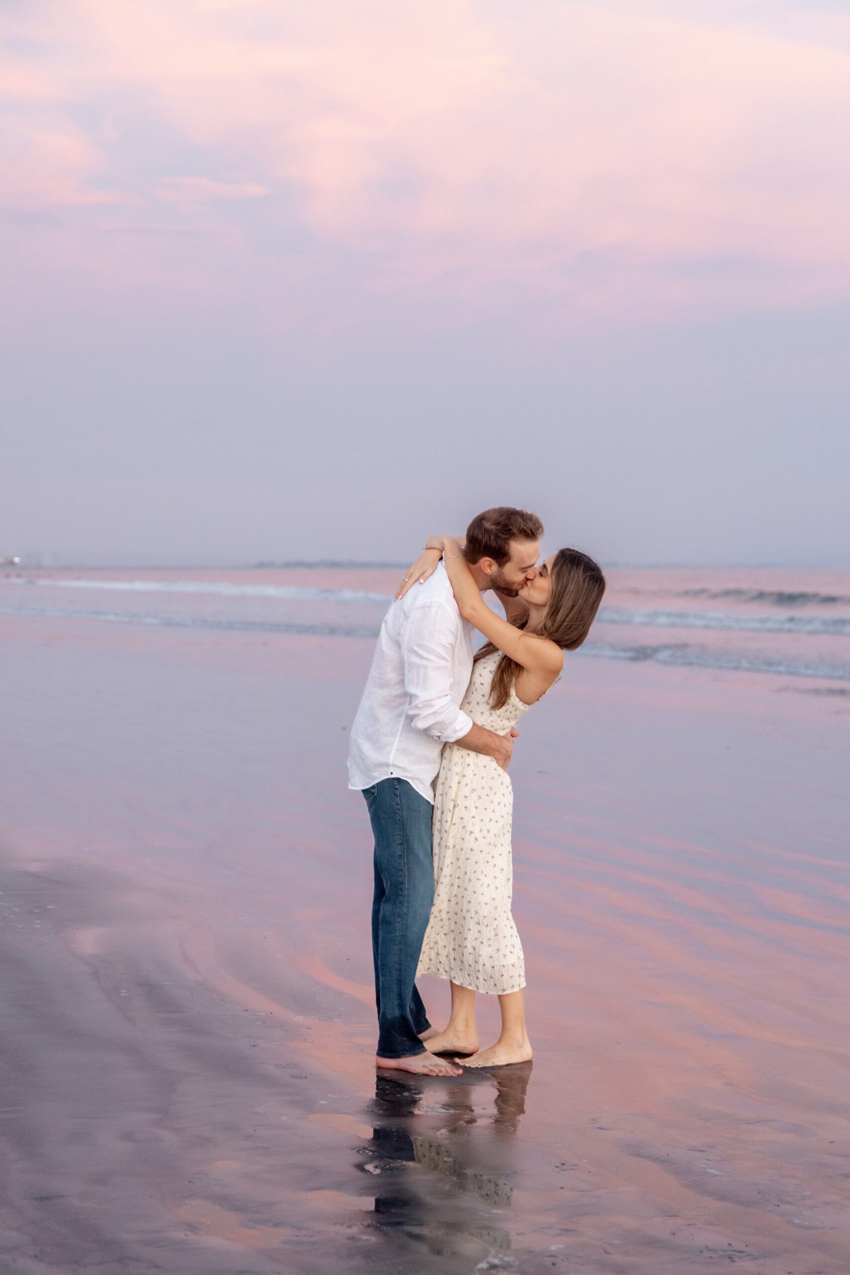 A happy engaged couple kisses on a beach under a beautiful pink sunset in a cream dress and white shirt with jeans