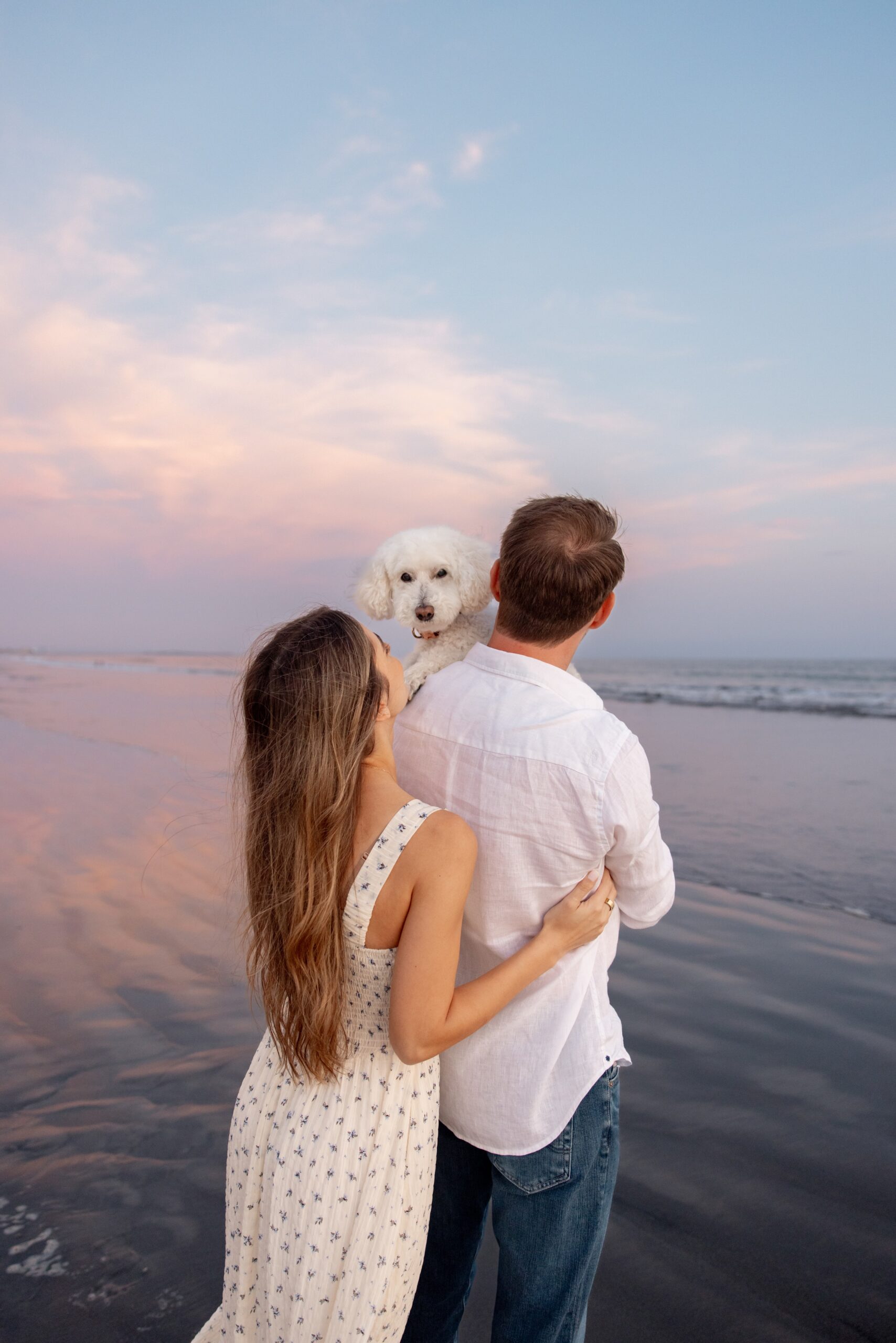 A woman looks up to her white dog on her fiancee's shoulder on a beach at sunset after visiting intimate wedding venues in san diego