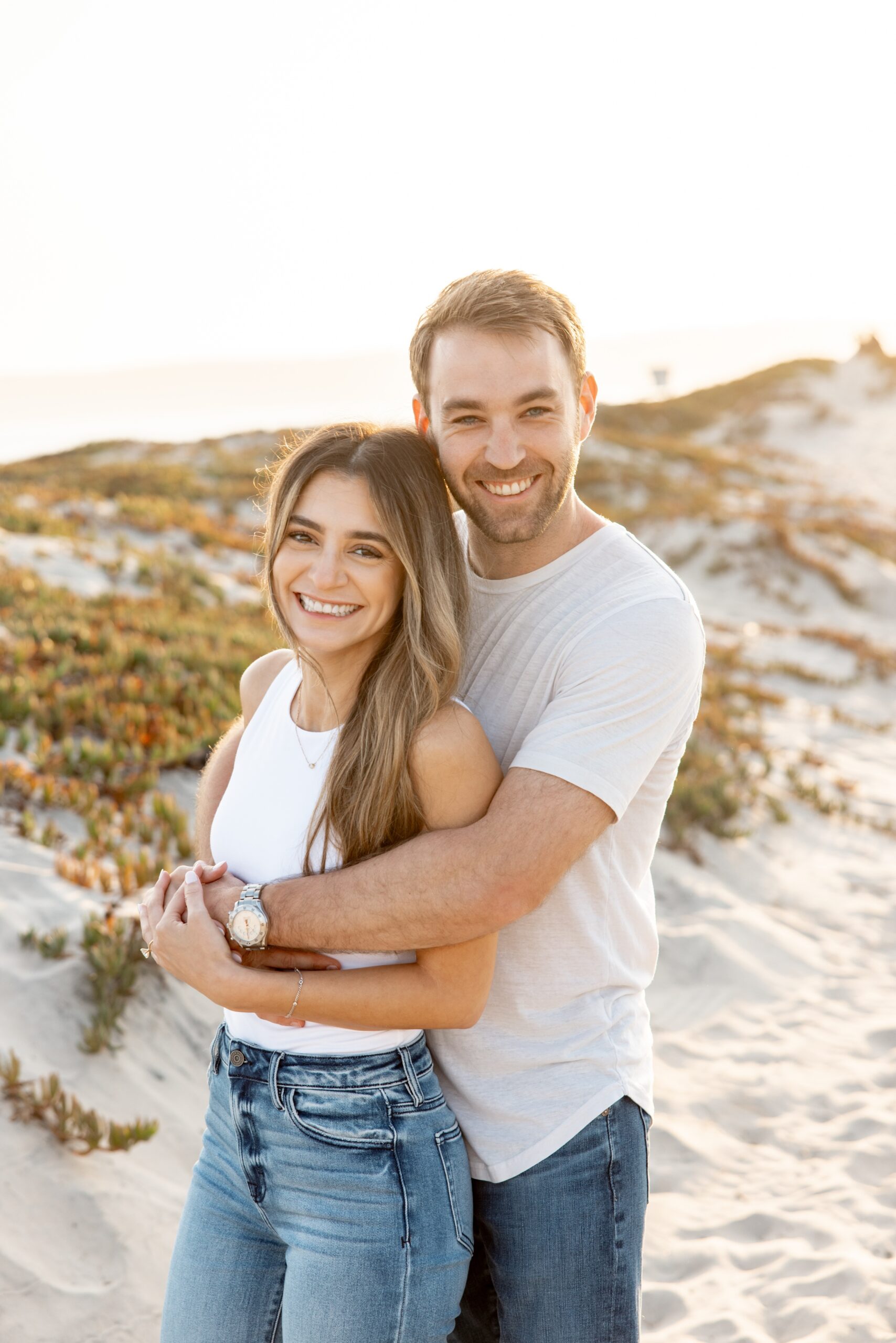 A happy engaged couple snuggle and laugh on a beach at sunset after visiting intimate wedding venues in san diego