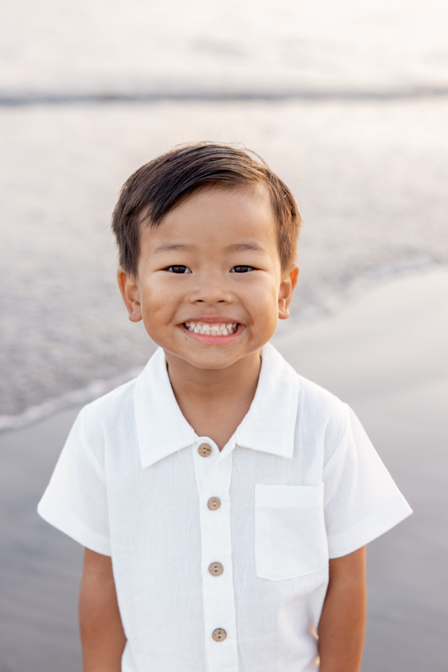 A happy young boy in a white shirt smiles while standing on a beach