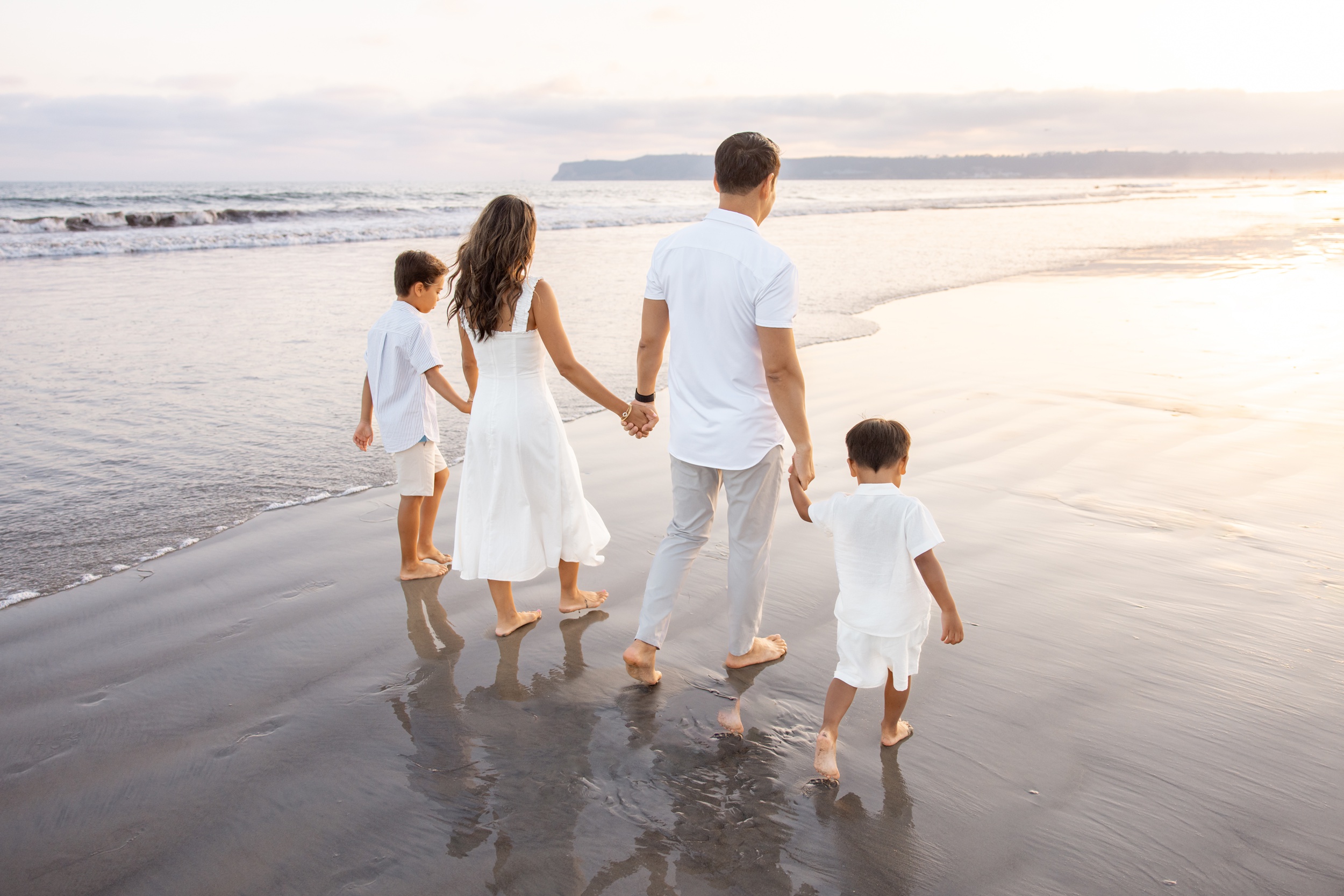 A mom and dad walk on a beach holding hands witht heir two young sons on a beach all in white before visiting san diego zoo with kids