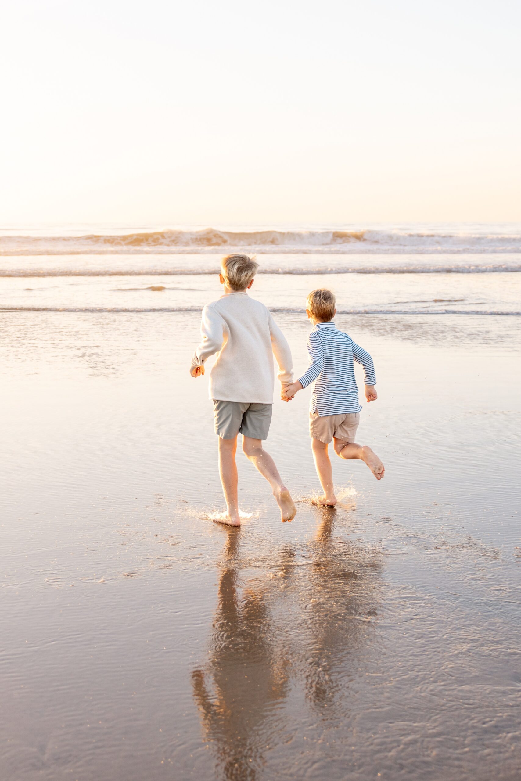 Two brothers in shorts and long sleeves run towards the water on a beach at sunset holding hands during summer activities in san diego