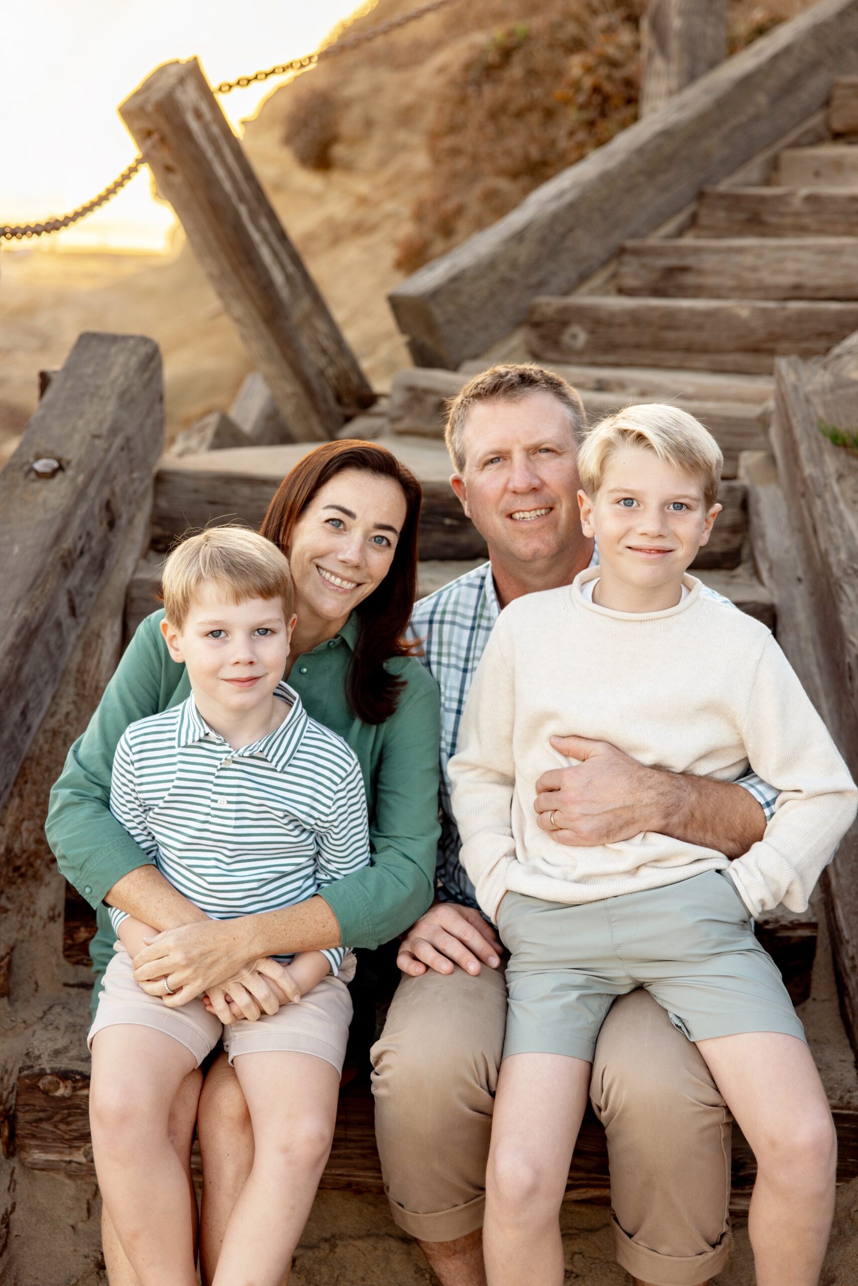 Happy mom and dad sit on the wooden beach stairs at sunset with their young sons in their laps during summer activities in san diego