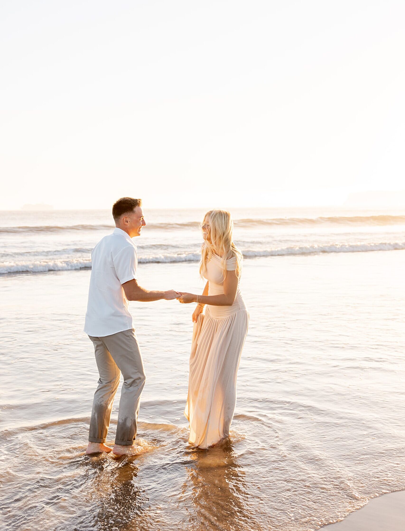 A happy engaged couple plays in the shallow water on the beach at sunset