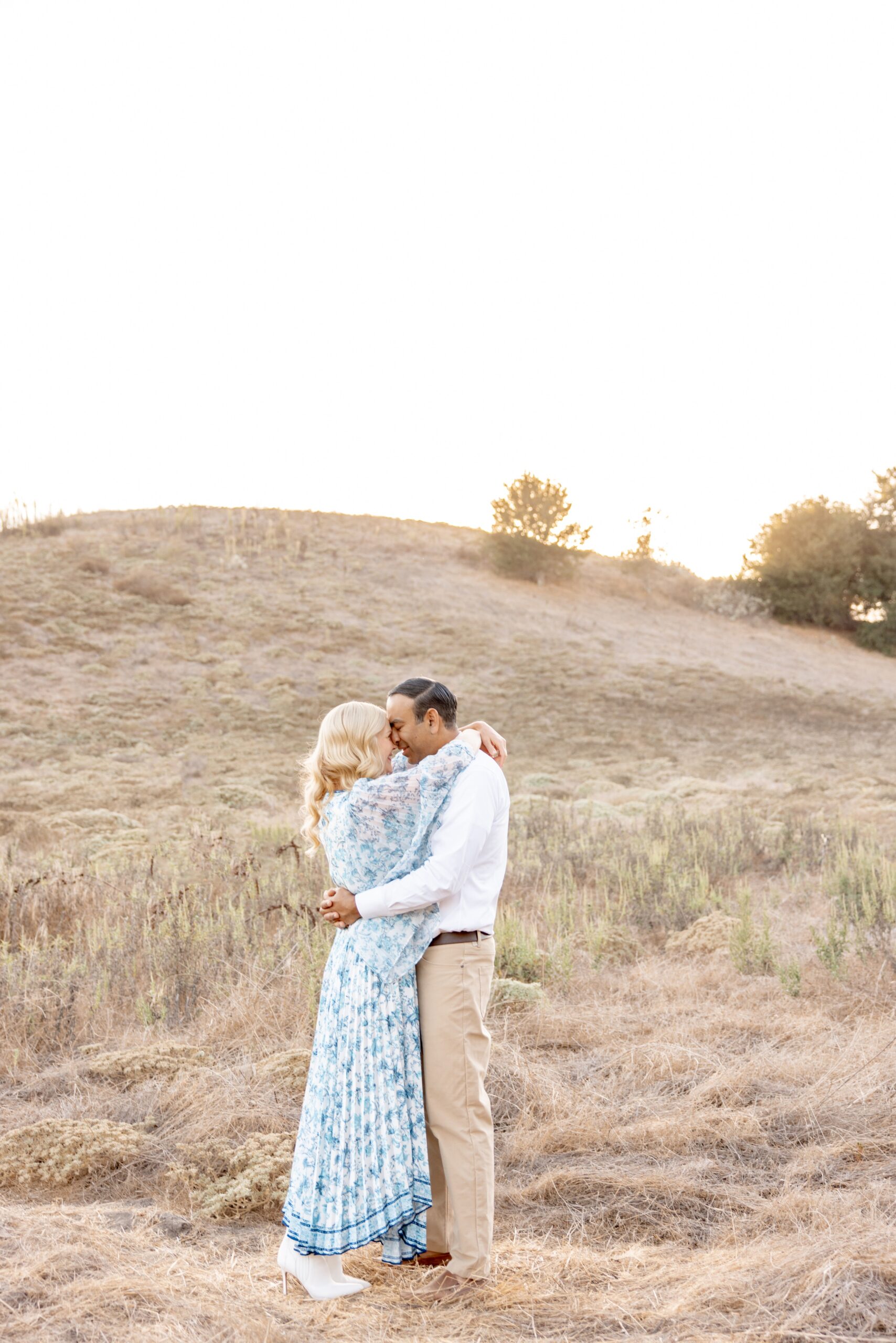 A happy couple hug and snuggle foreheads in a park hillside in a blue dress and khakis before a sunset cruise in san diego