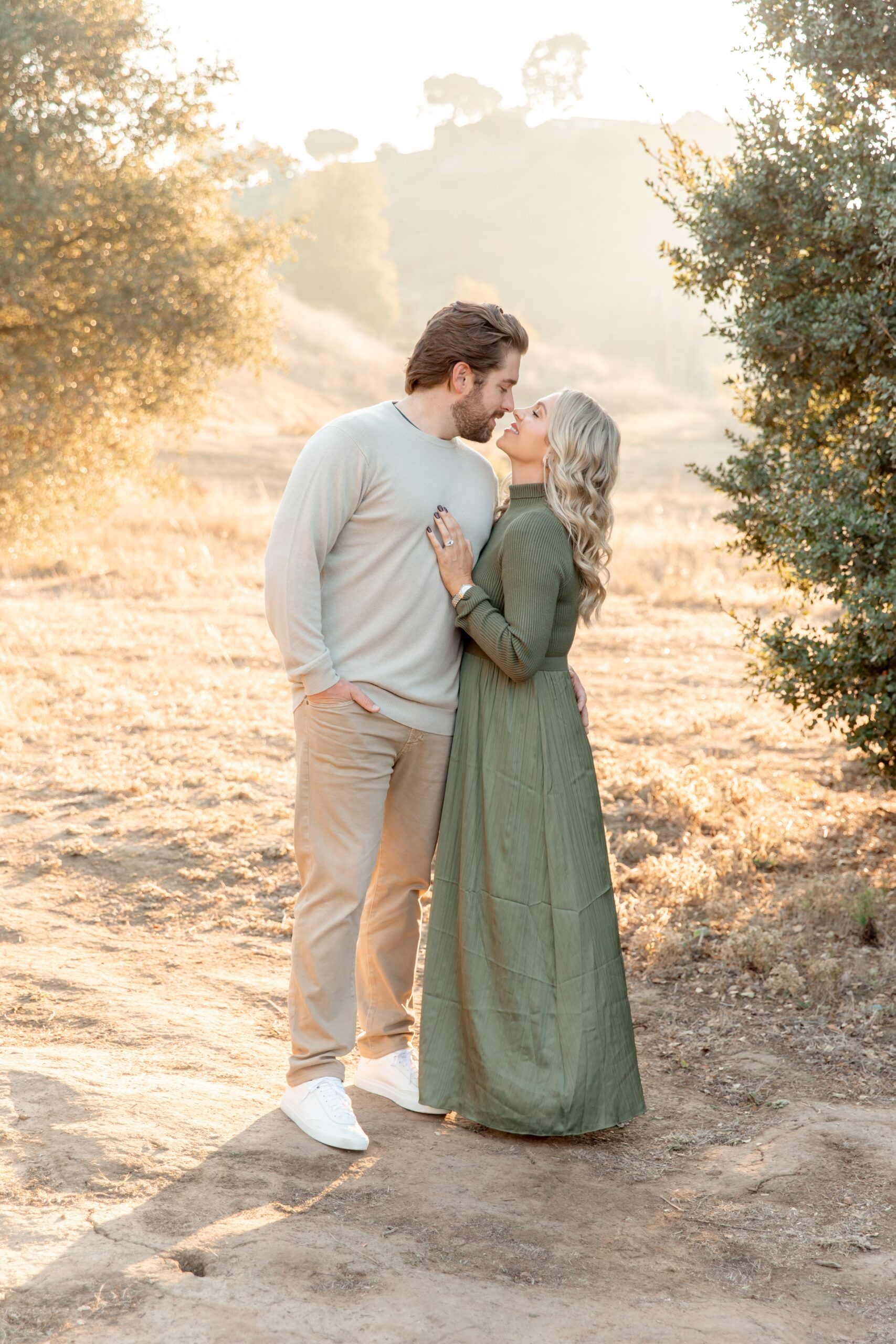 A happy couple in green and beige lean in for a kiss in a park at sunset before a sunset cruise in san diego