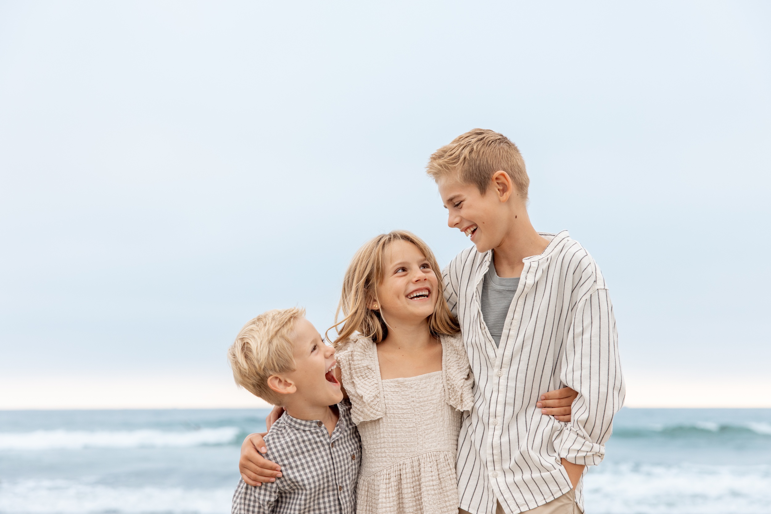 Three young siblings laugh on a beach with arms around each other