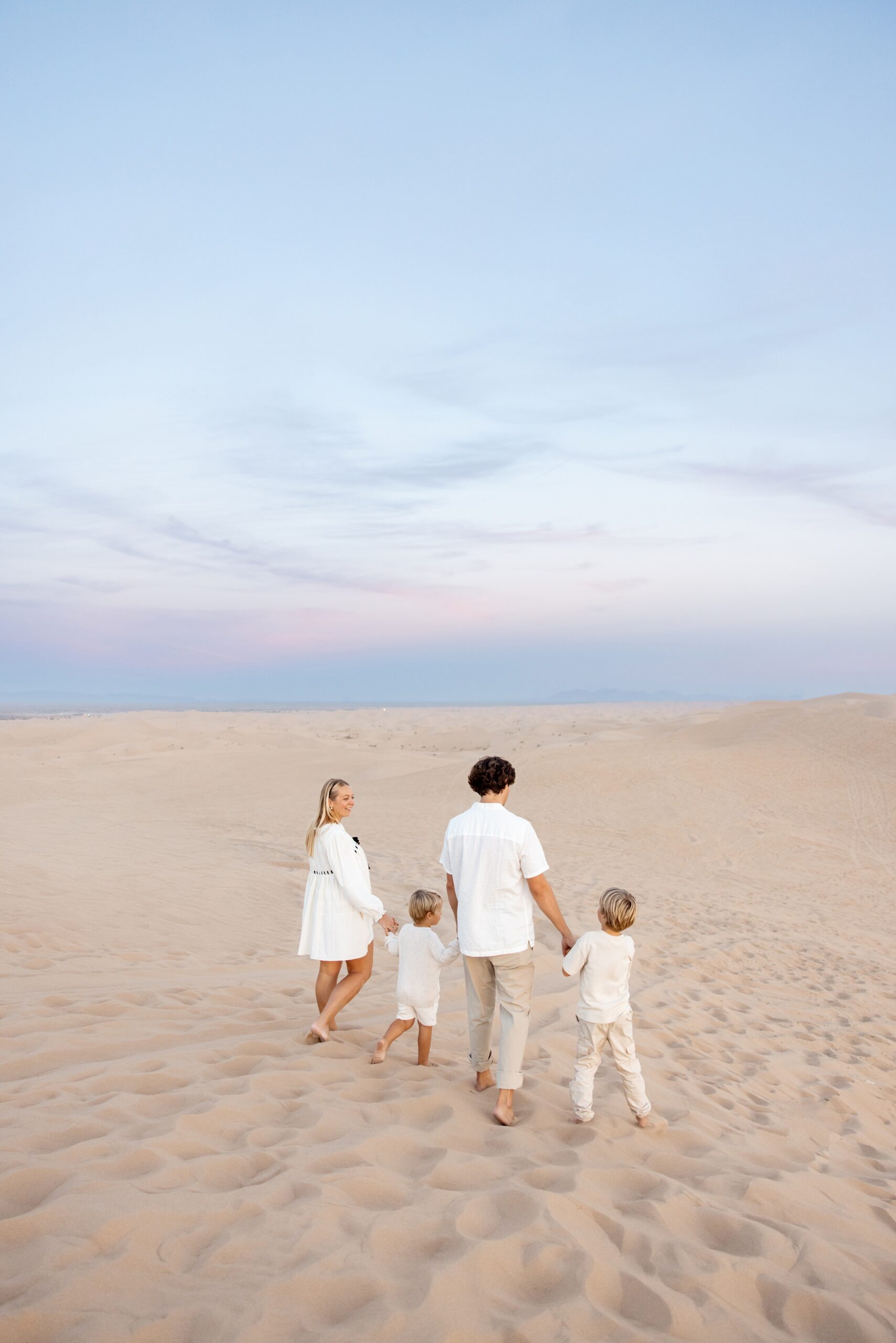 A happy family walks on a sand dune in all white holding hands and laughing before visiting trampoline parks in san diego