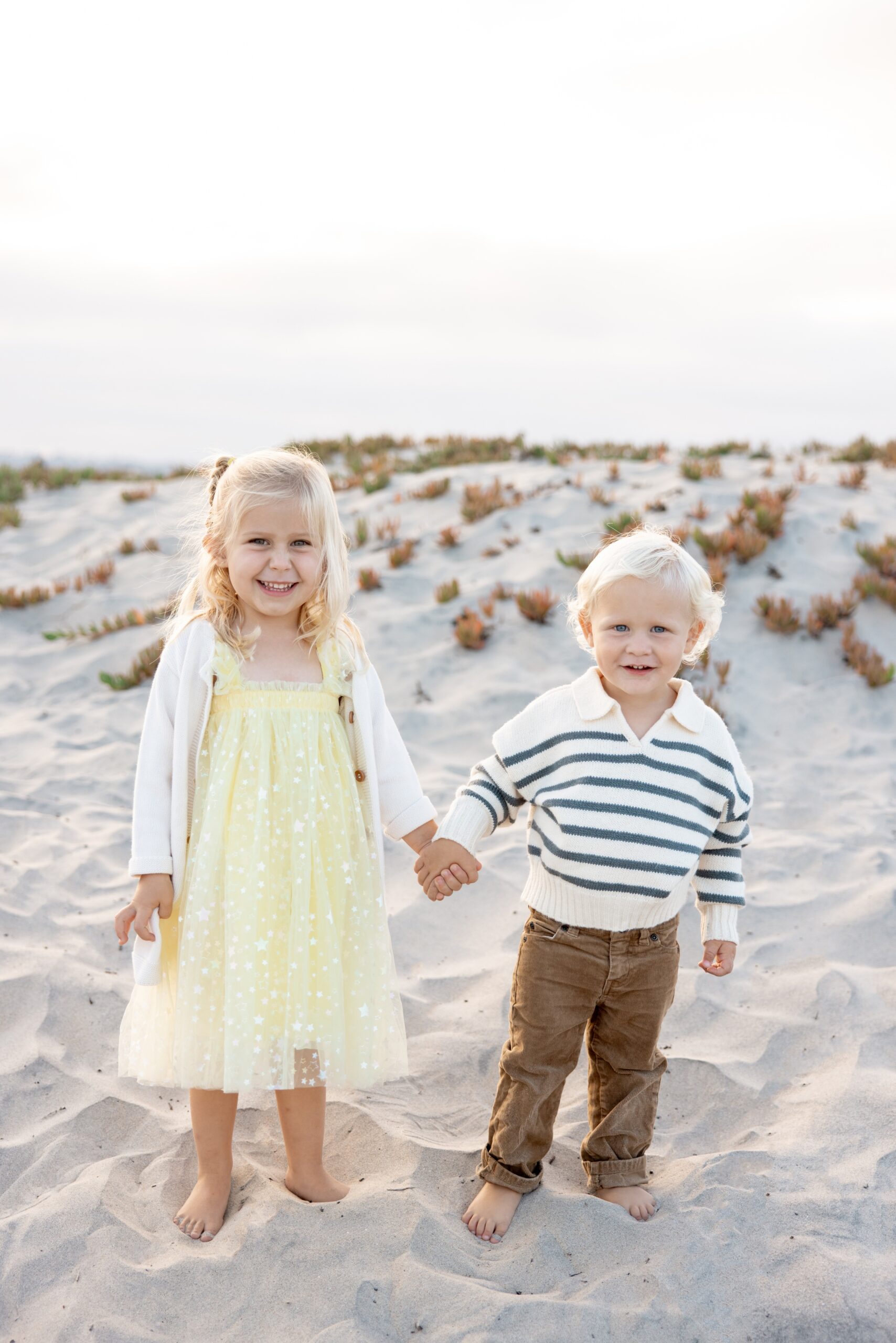 A toddler girl in a yellow dress smiles while holding hands with her younger brother on a beach before visiting trampoline parks in san diego