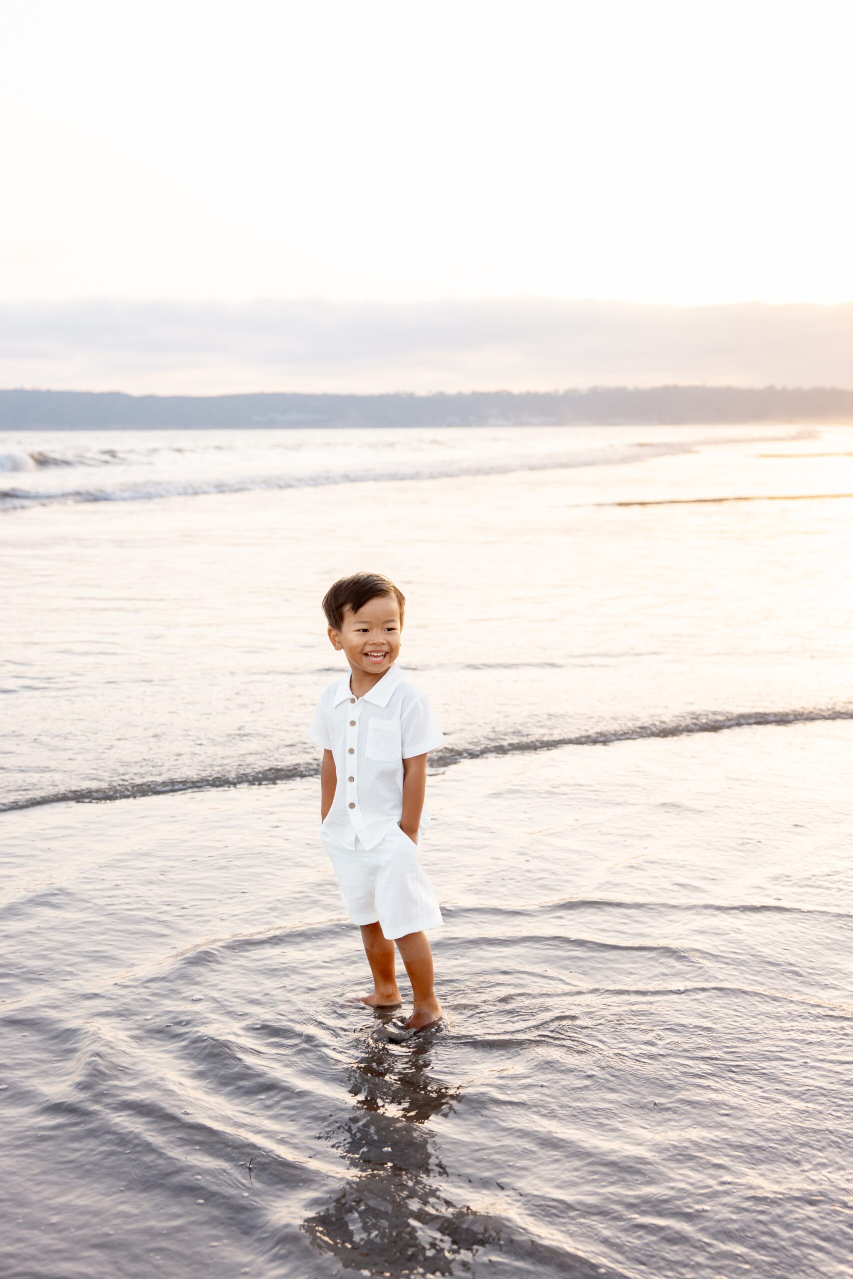 A happy young toddler boy in all white stands in the water with hands in his pockets on a beach at sunset