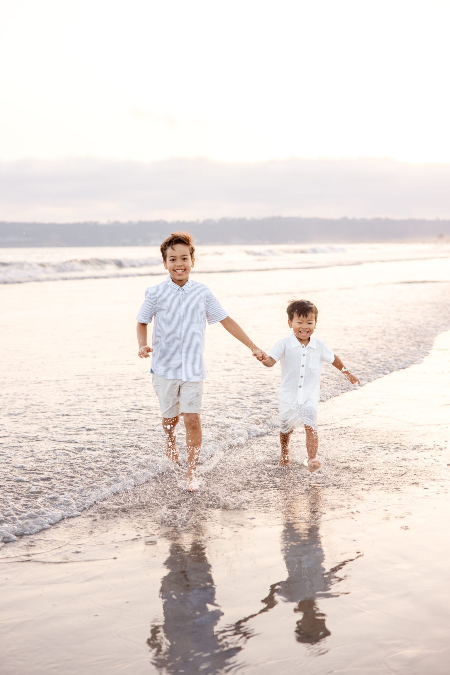Two brothers run through the water on a beach holding hands after visiting waterparks in San Diego