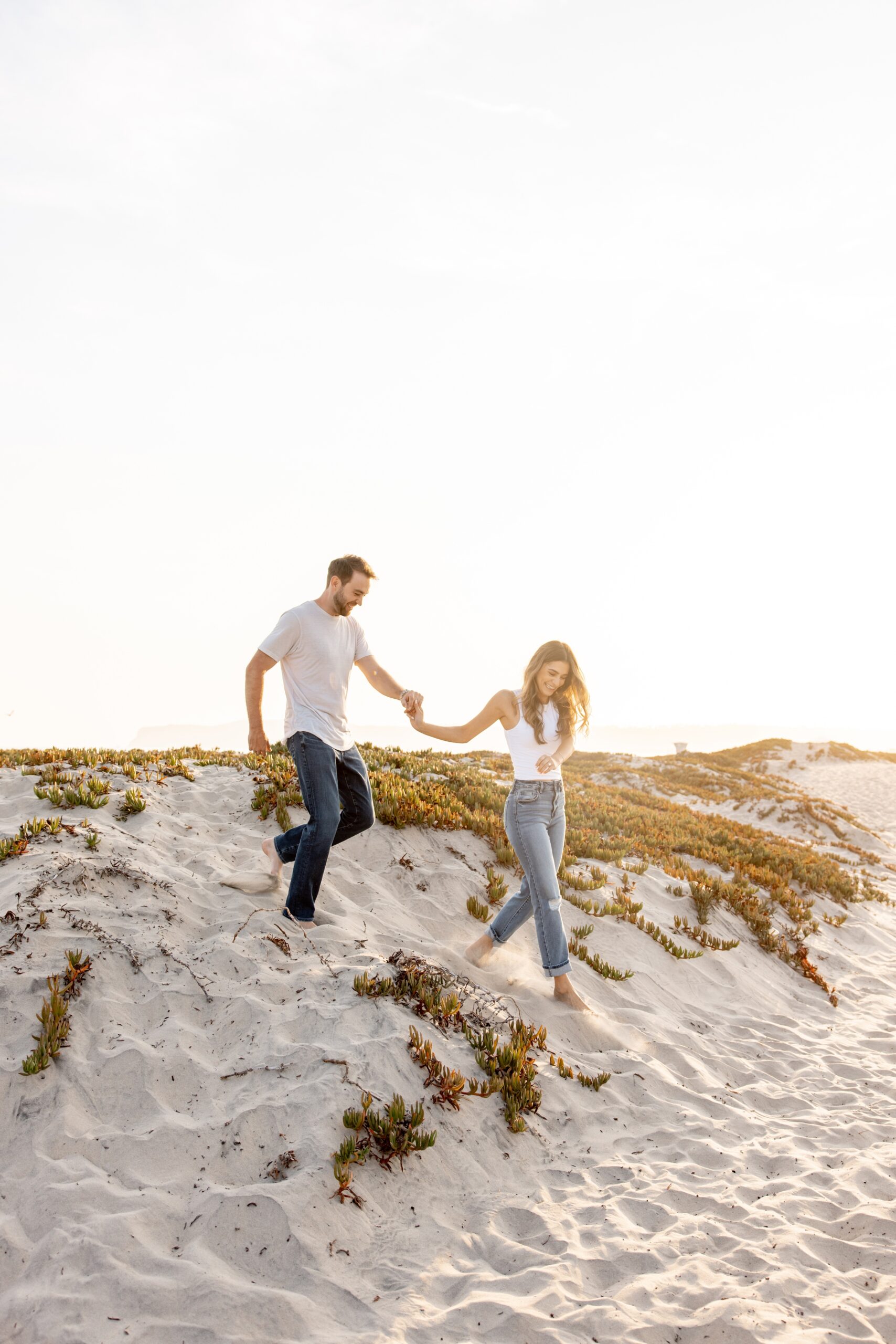 A woman in a white shirt and jeans leads her fiancee down a beach dune by the hand at sunset after exploring where to propose in san diego