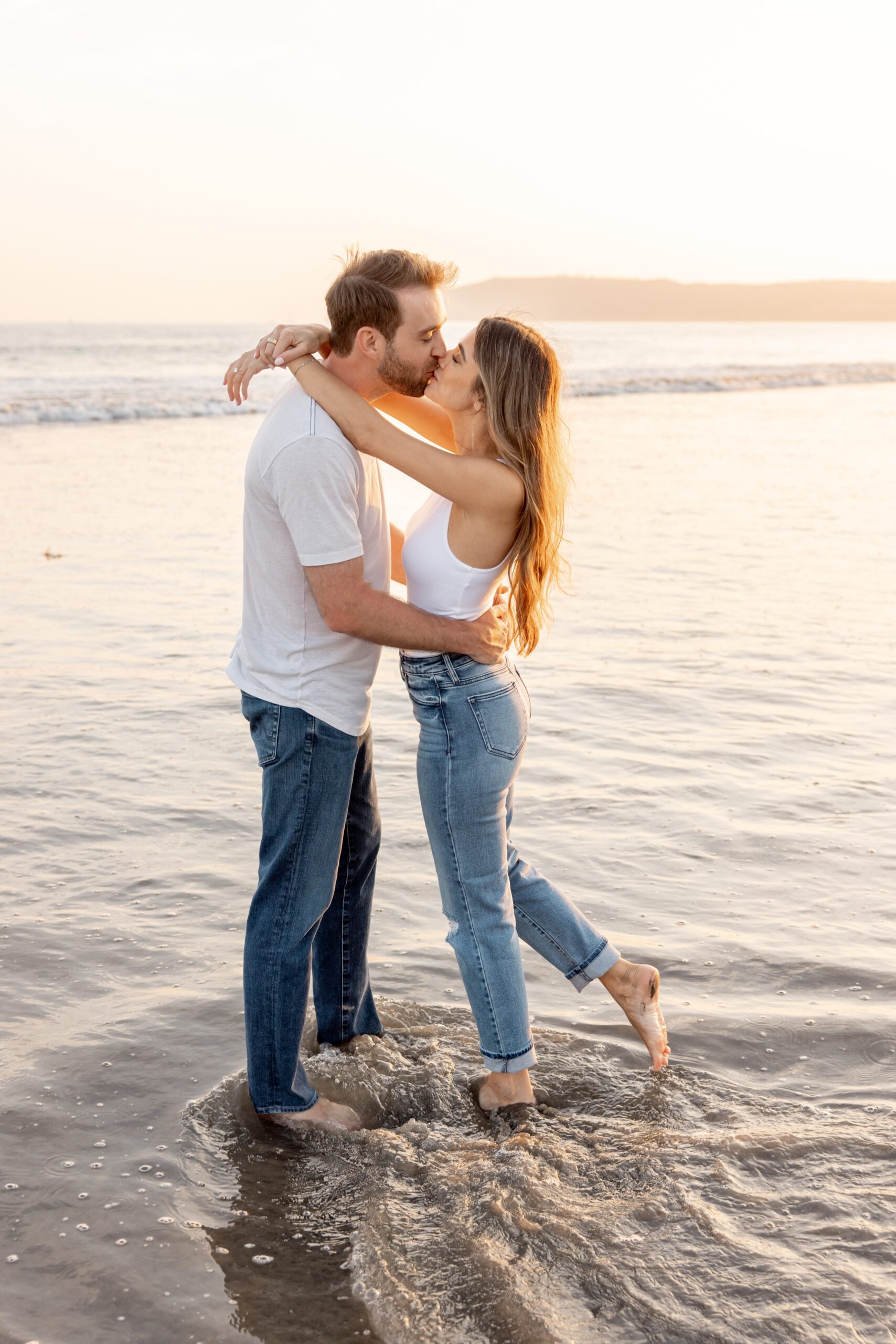 A happy engaged couple in white shirts and jeans hug and kiss on a beach at sunset with feet in the water after finding where to propose in san diego