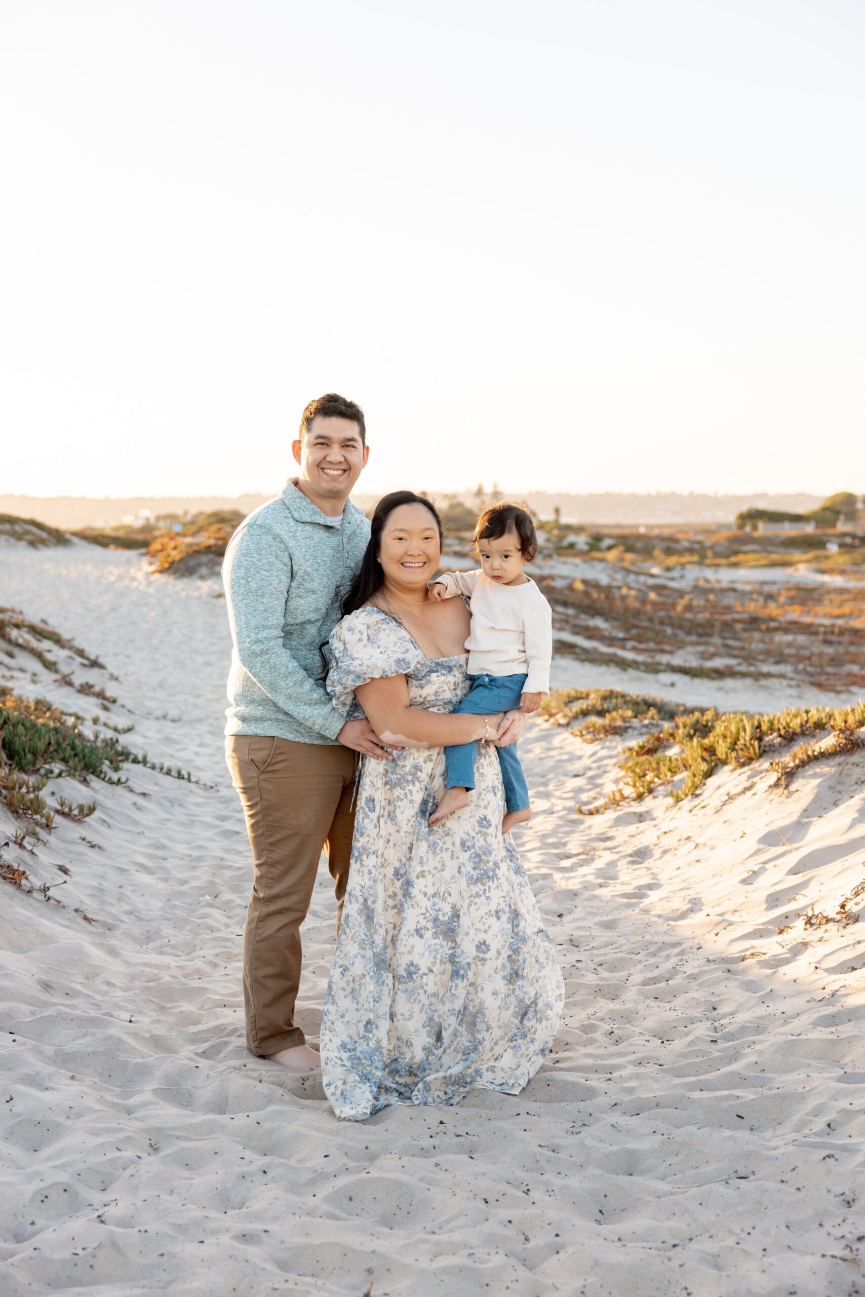 A mom and dad smile while standing on a beach dune holding their toddler before exploring 4th of July events in San Diego