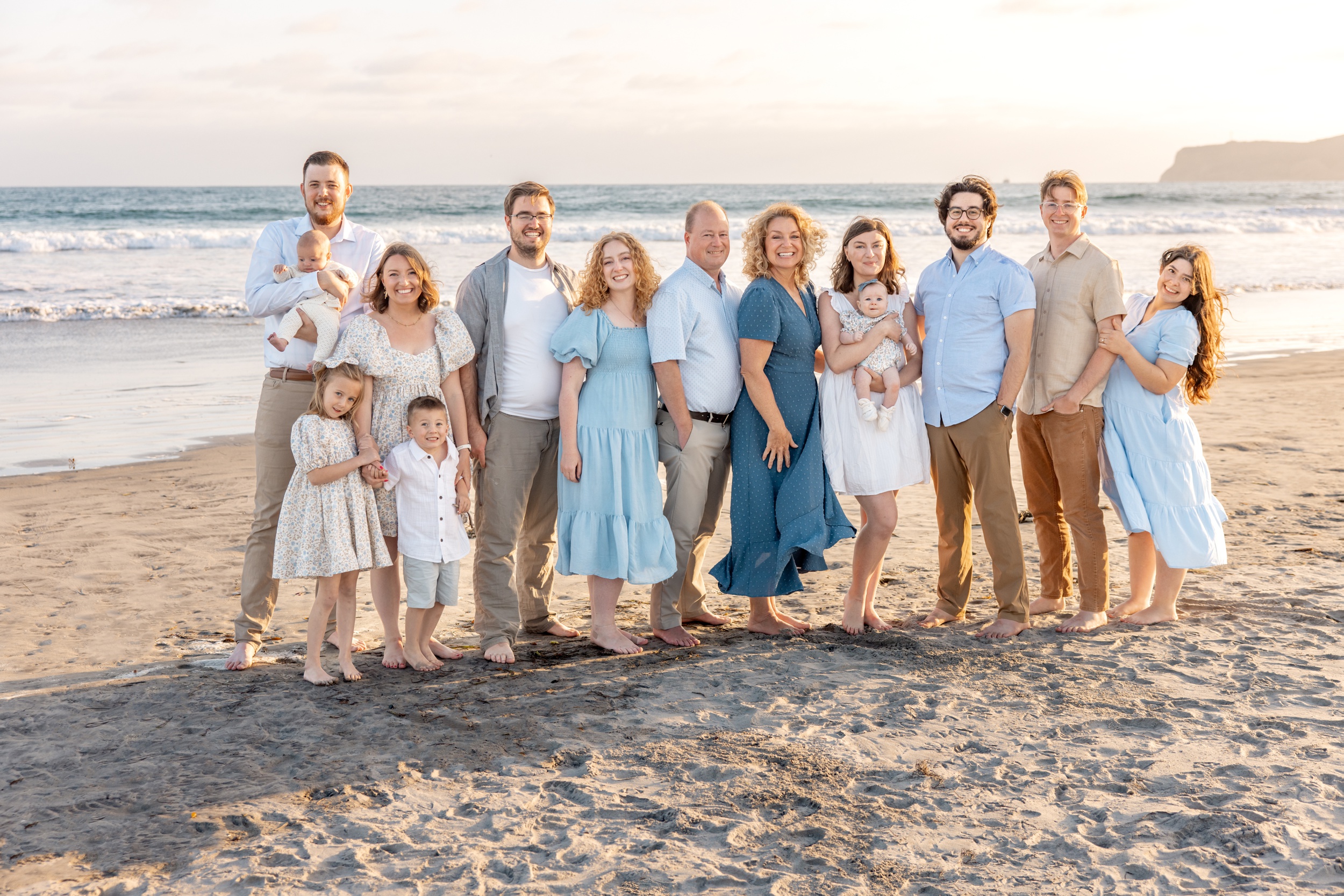 A happy large extended family stands together with three generations on a beach smiling