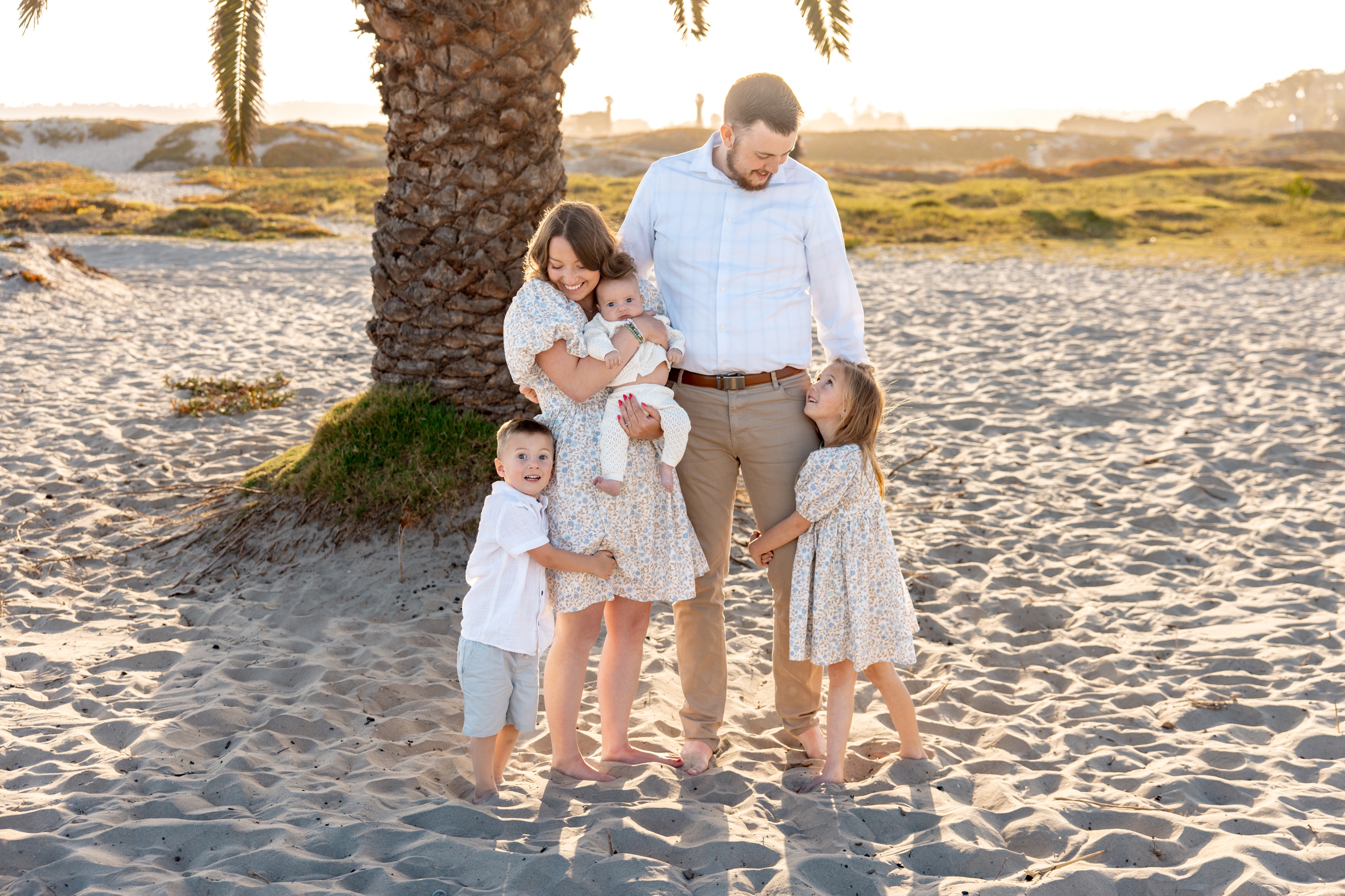 Toddler brother and sister hug mom and dad's legs while standing on a beach with infant in mom's arms