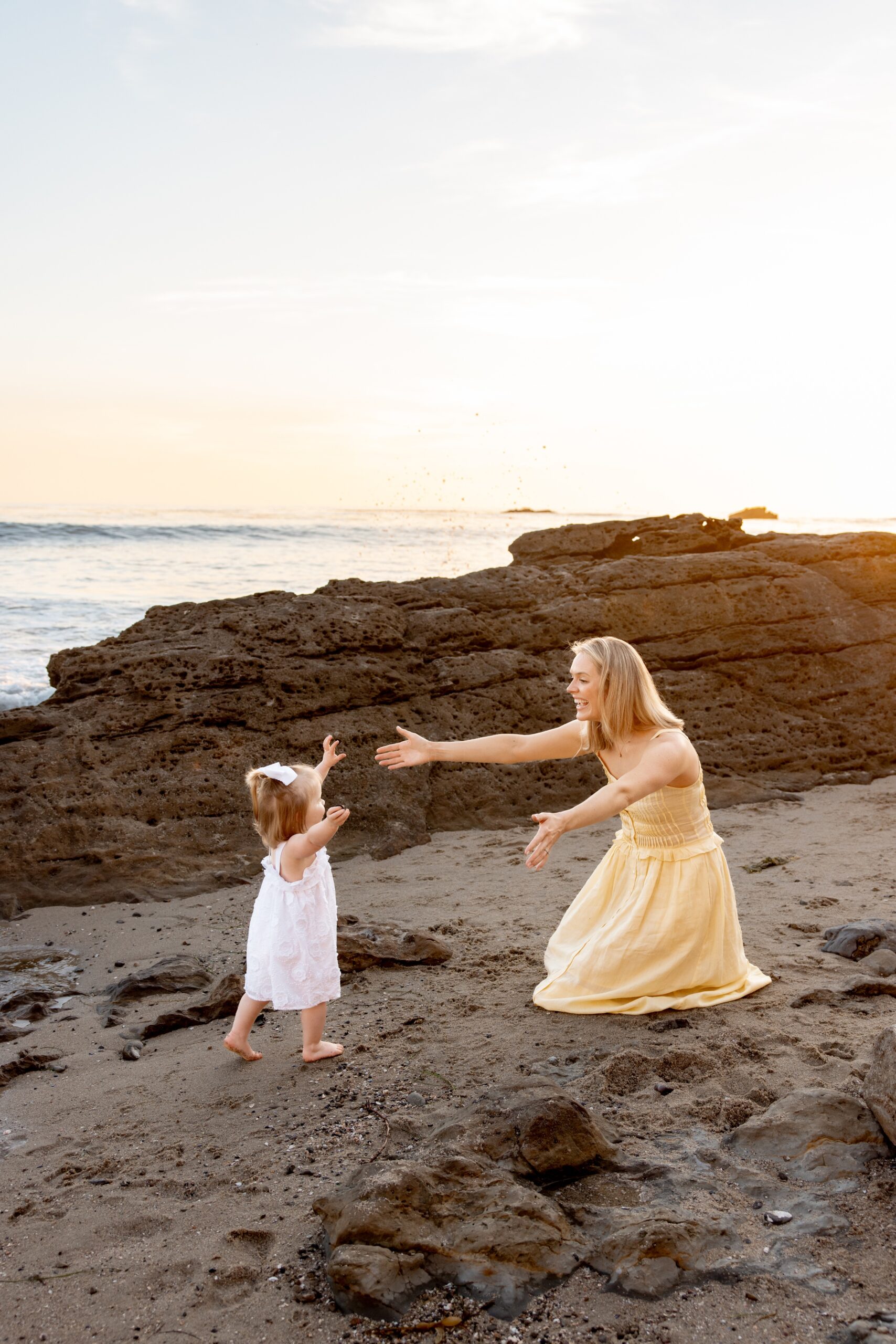 A mom kneels with arms out on a beach as her toddler daughter runs towards her for a hug