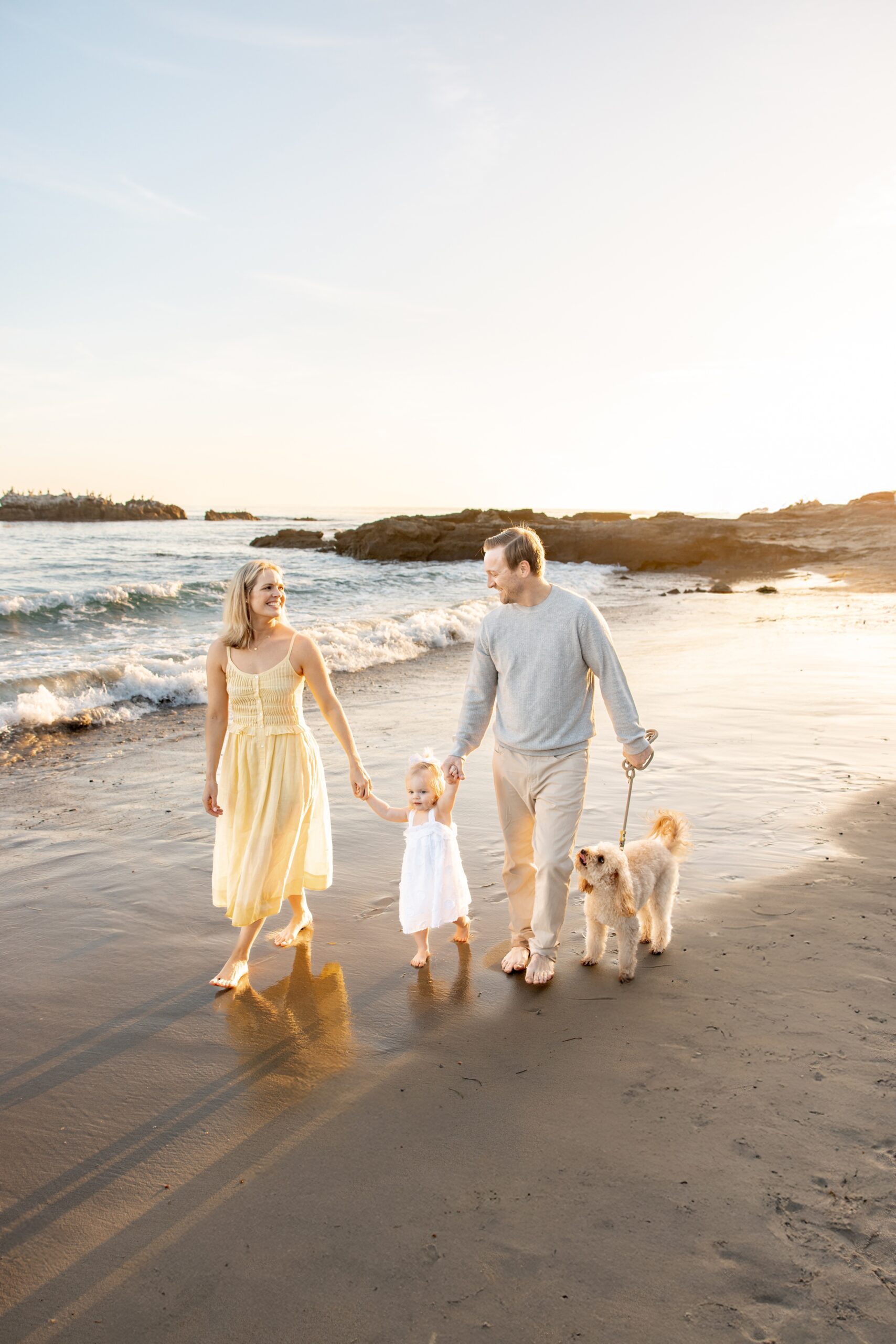 A happy mom and dad walk on a beach holding hands with their toddler daughter and walking their small blonde dog after visiting geppetto's toys