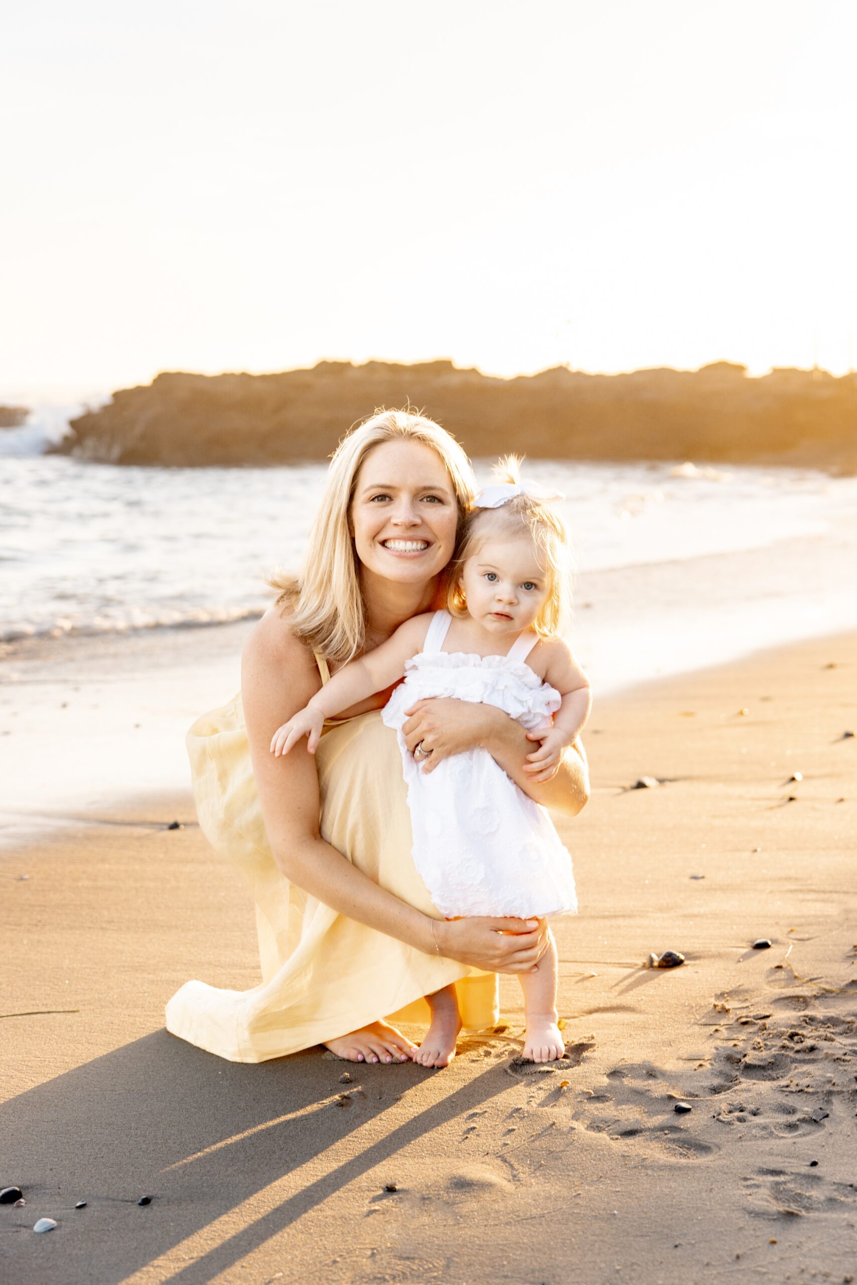 A happy mom in a yellow dress kneels to hug her toddler daughter on a beach after visiting geppetto's toys