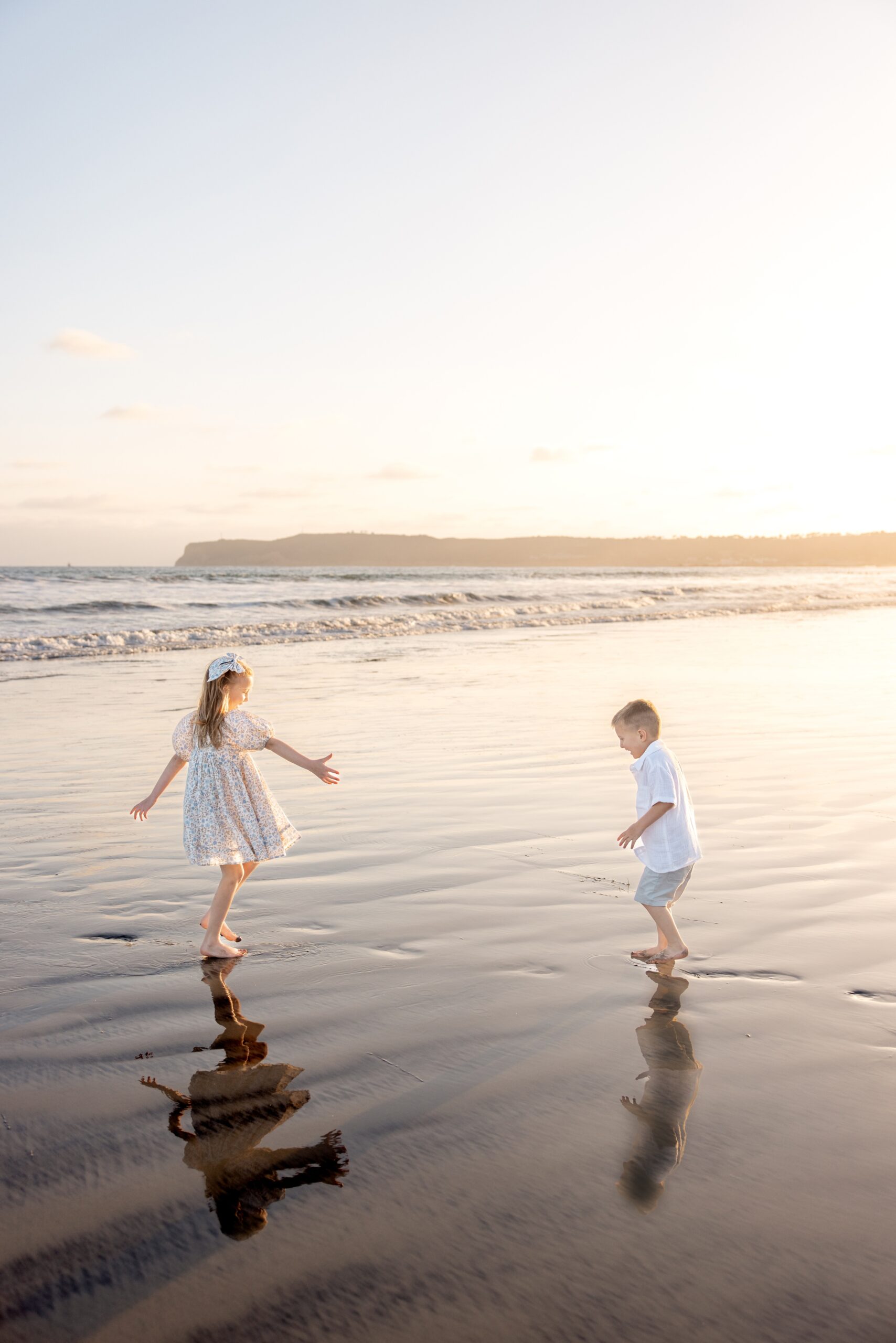 Toddler brother and sister play in the low tide on a beach at sunset in white outfits