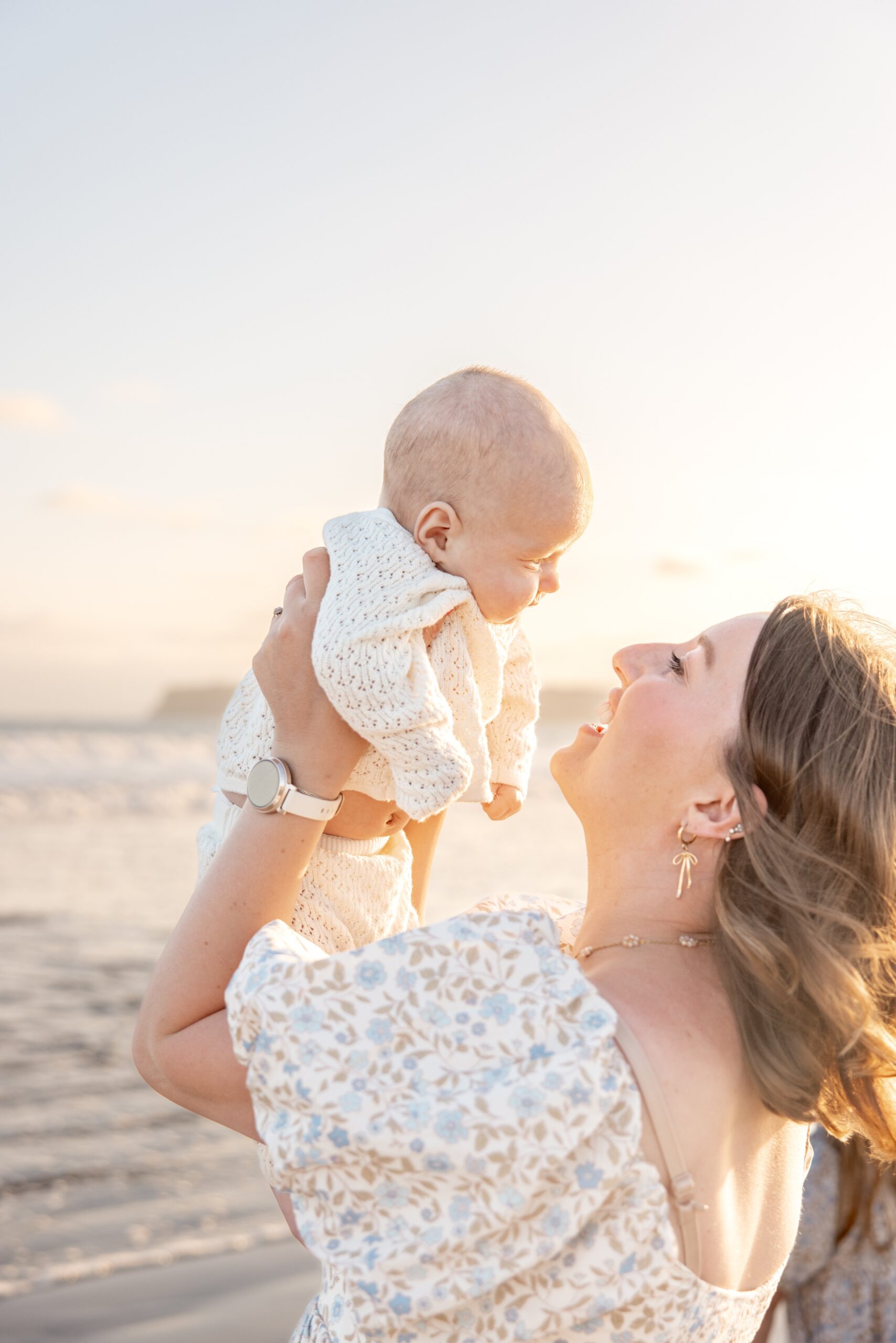A happy mom lifts her infant in a white sweater above her head on a windy beach at sunset after visiting Padres Kidfest