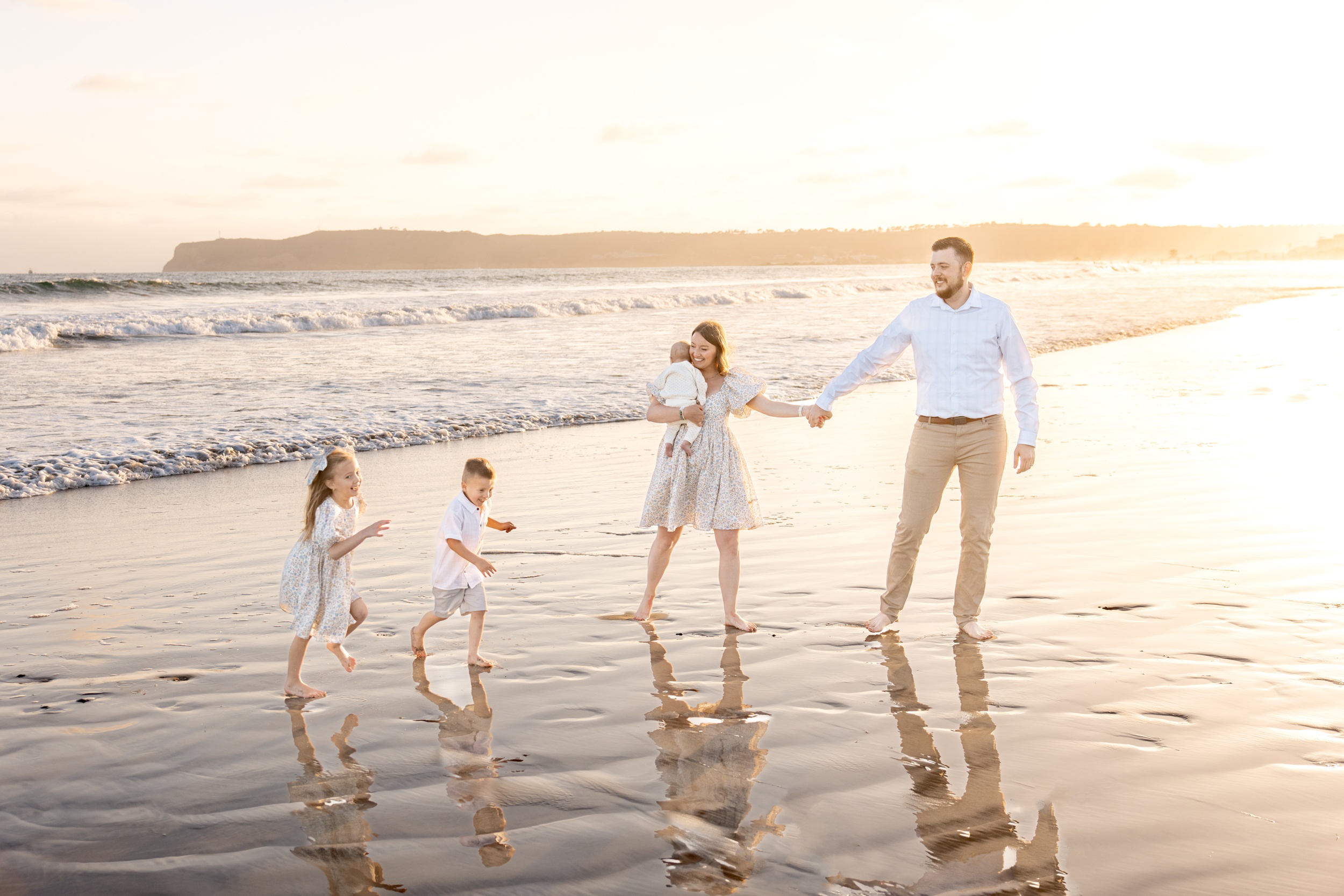 A mom and dad hold hands on a beach while their two toddlers run and laugh at sunset before heading to Padres Kidfest