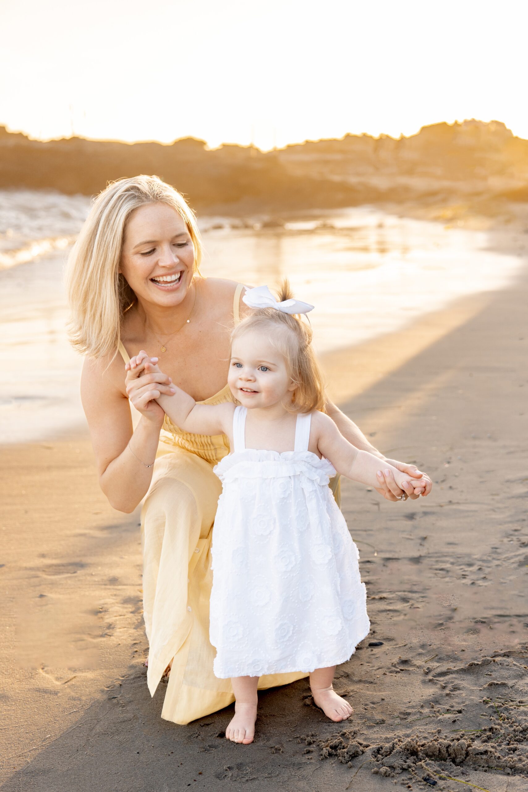 A happy mom plays with her daughter on a beach in white and yellow dresses
