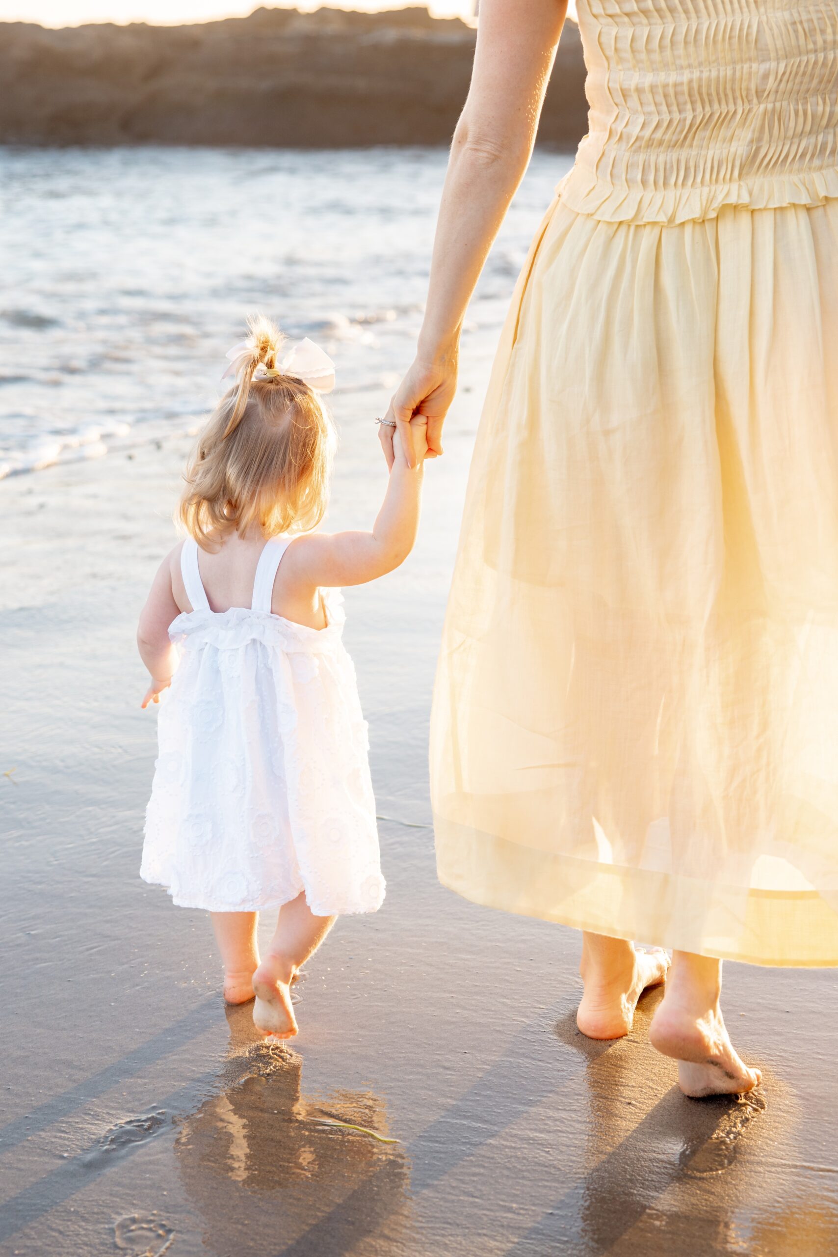 A mom in a yellow dress walks her toddler daughter in a white dress on a beach at sunset after visiting play city san diego
