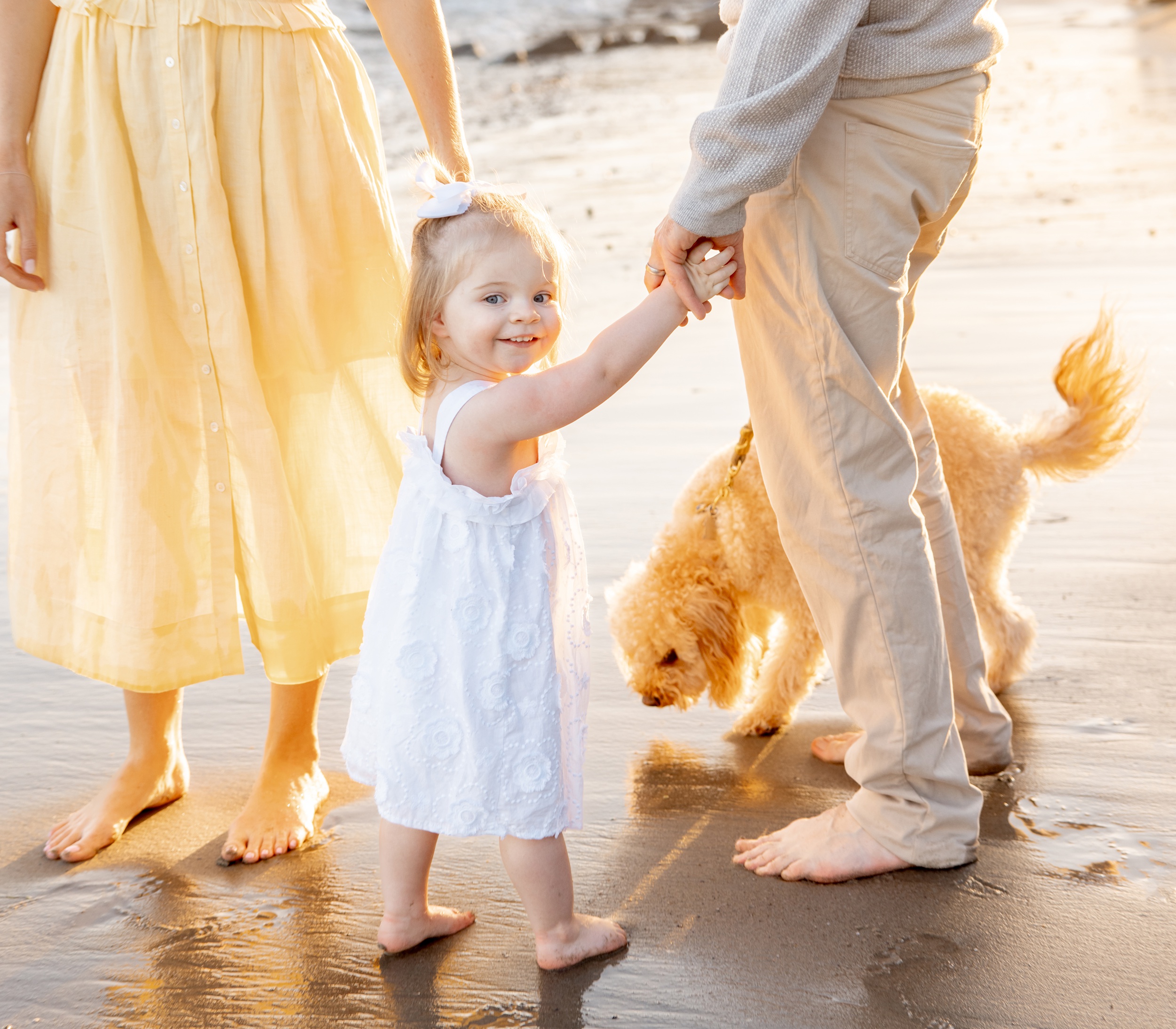 A happy toddler girl walks on a beach smiling over her shoulder and holding mom and dad's hands before visiting play city san diego
