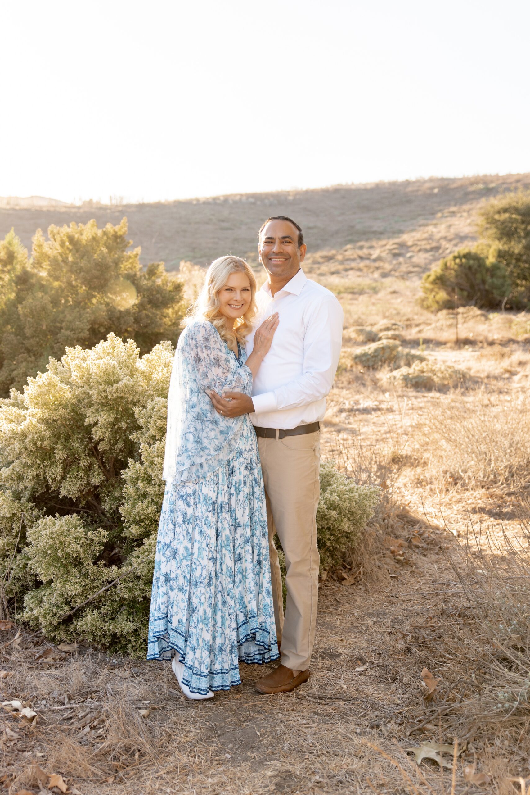 A happy couple smiles while standing in a desert park at sunset in a blue dress and white shirt