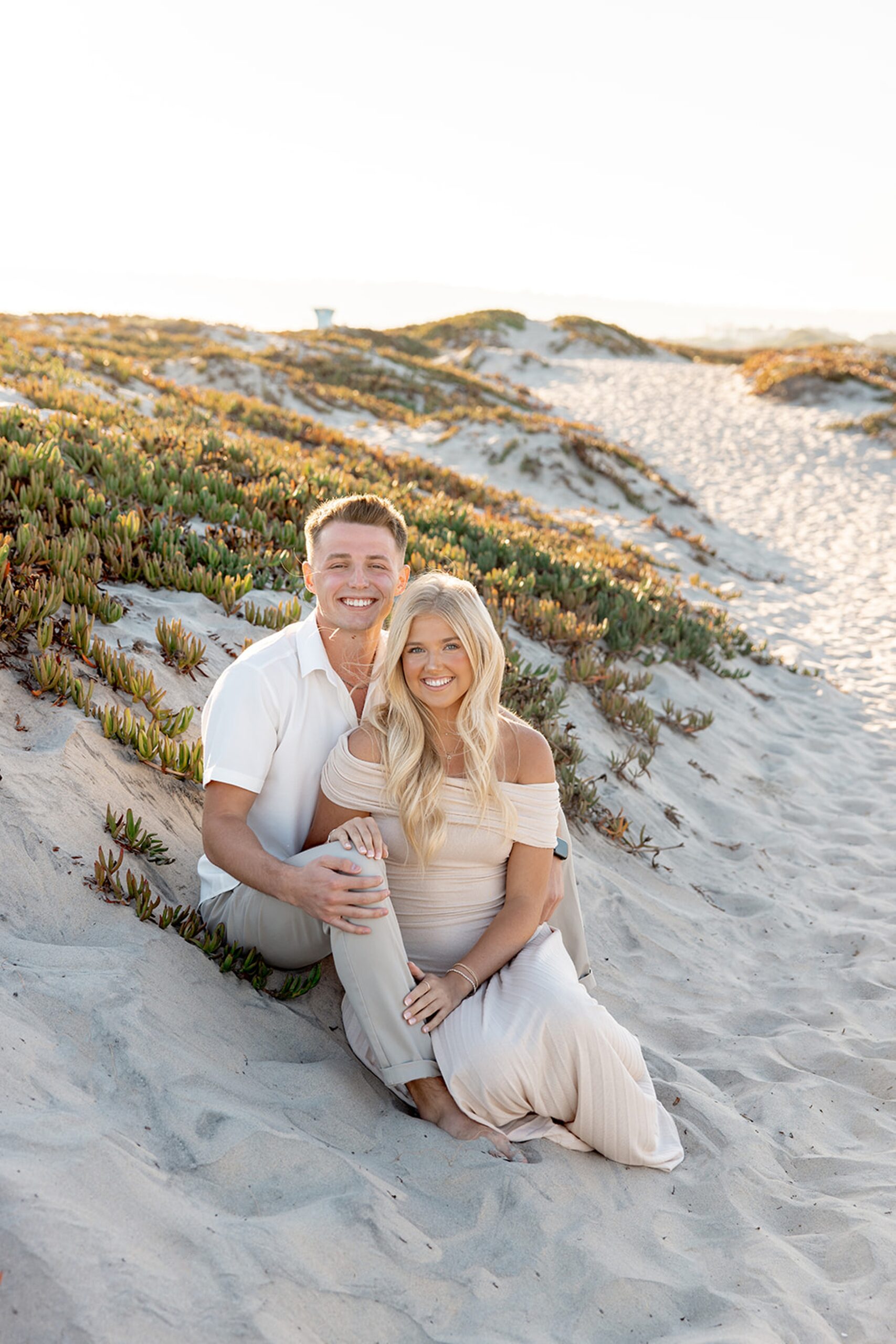 A woman in a cream gown leans into the lap of her partner on a beach at sunset after visiting the San Diego County Fair