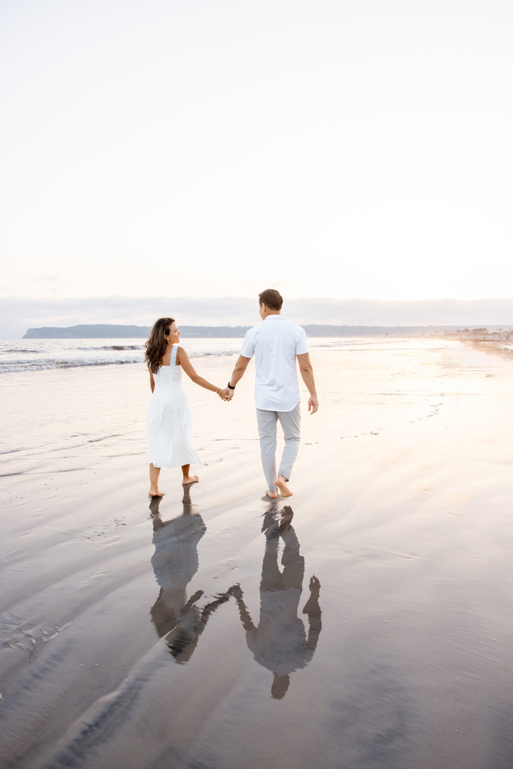 A happy couple wearing white walk hand in hand up a beach at sunset after visiting the San Diego County Fair
