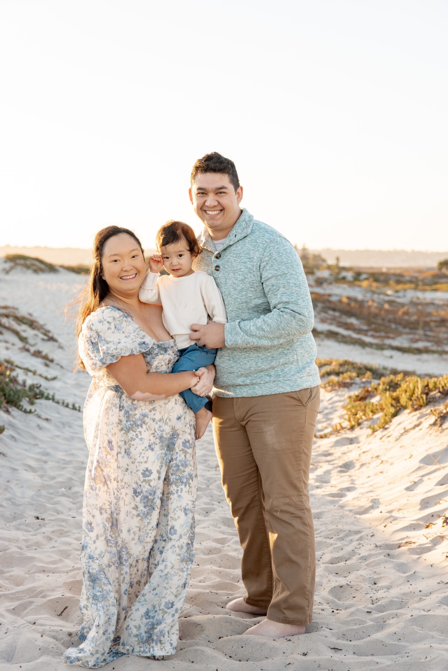 Happy mom and dad smile while holding their infant and standing on a beach dune