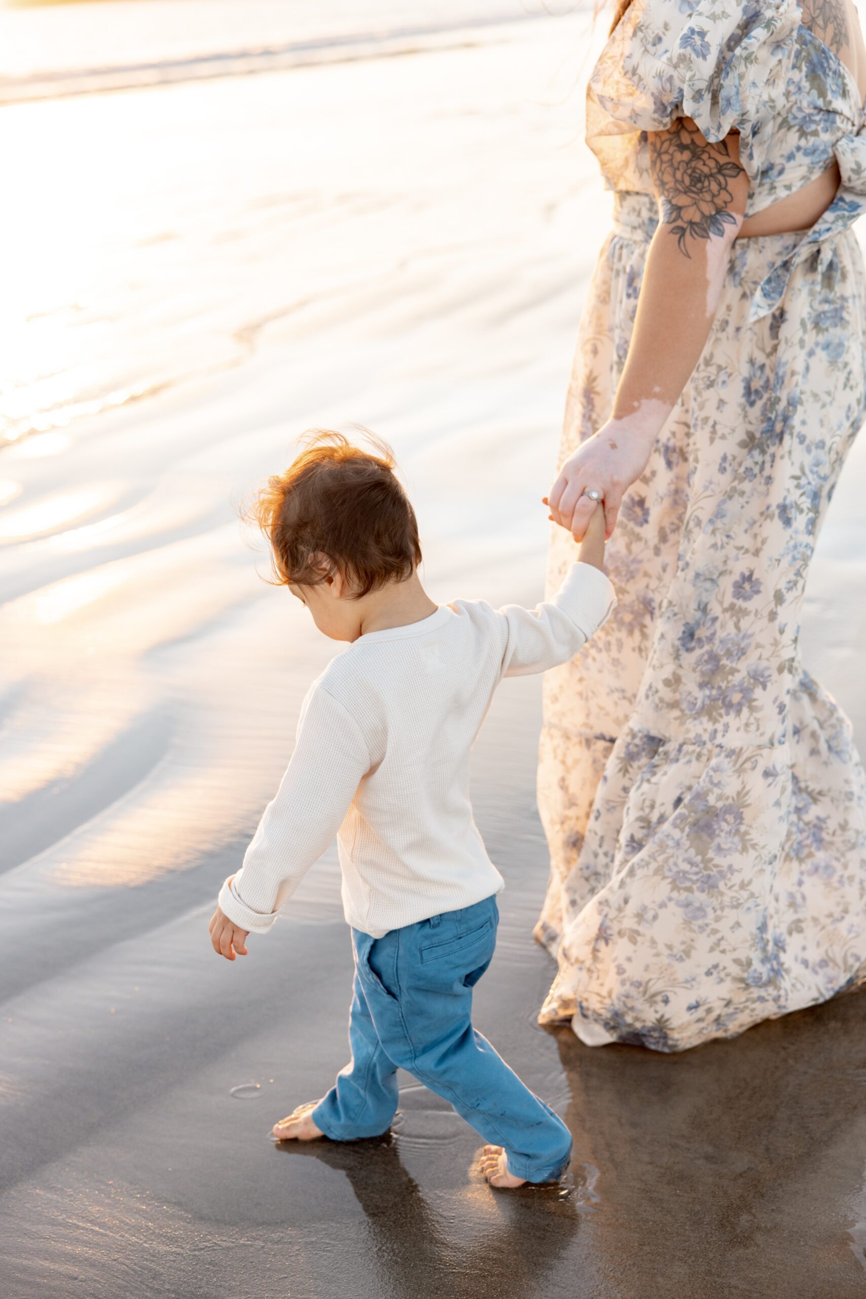 A mother in a floral print dress walks her toddler in blue pants and white shirt on a beach at sunset after visiting Sesame Place San Diego