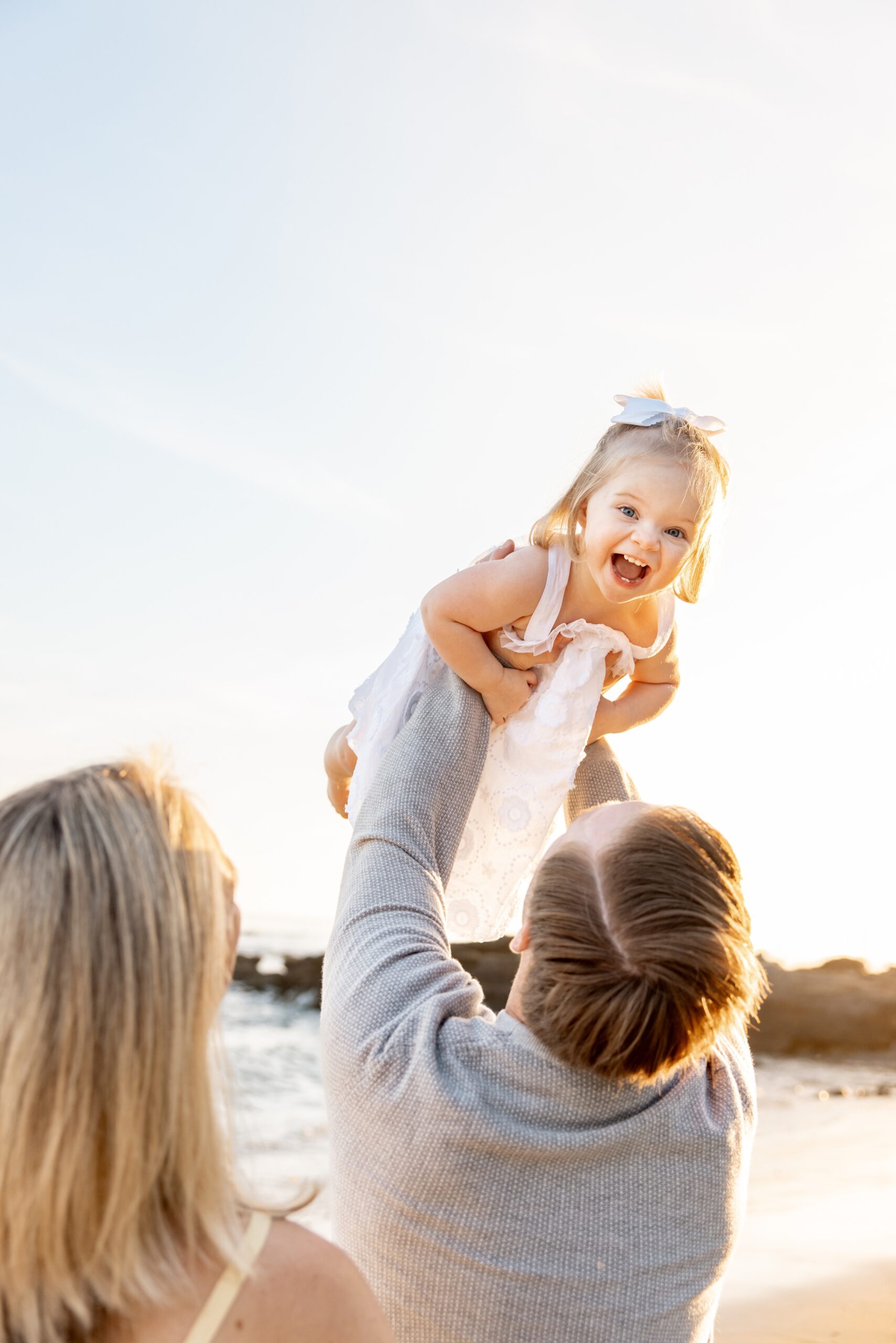 A dad in a grey sweater lifts his giggling toddler daughter above his head on a beach after enrolling in summer reading program in san diego