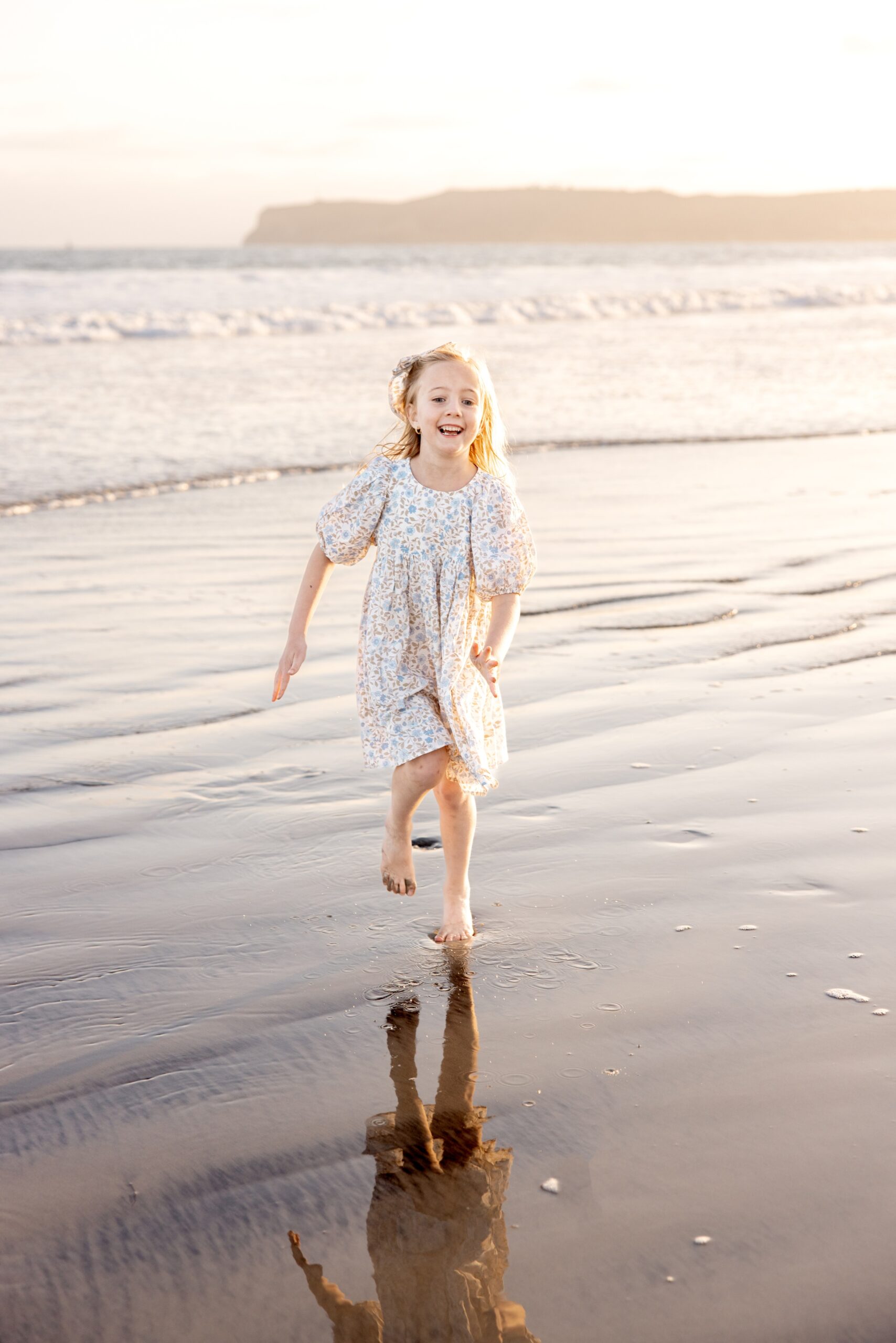 A giggling toddler girl in a floral print dress runs on the beach at low tide at sunset