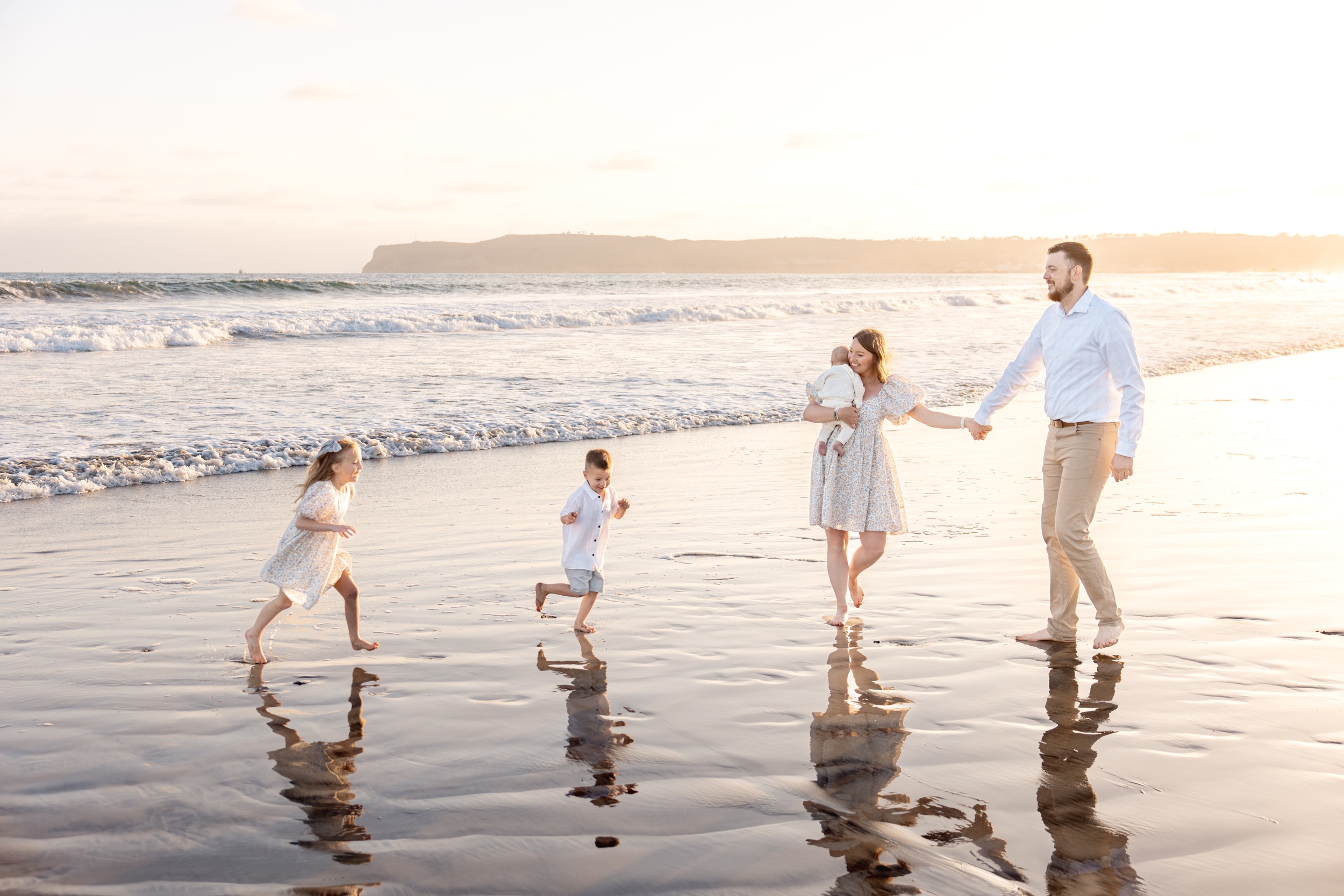 A mom and dad walk on a beach holding hands and infant as their two toddlers run around at sunset before visiting Birch Aquarium exhibits