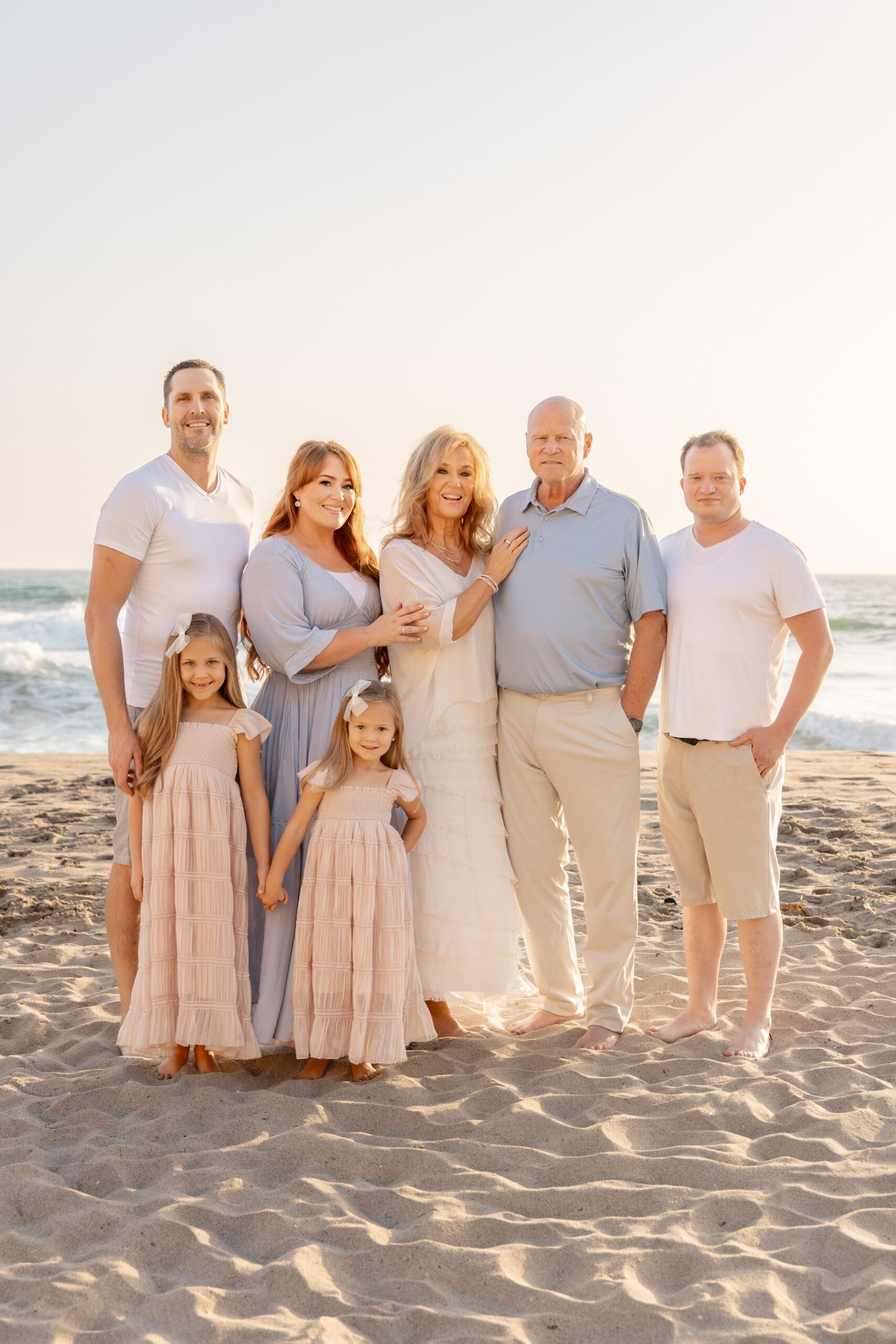 An extended family with two toddler girls stand on a beach at sunset smiling after visiting country clubs in san diego