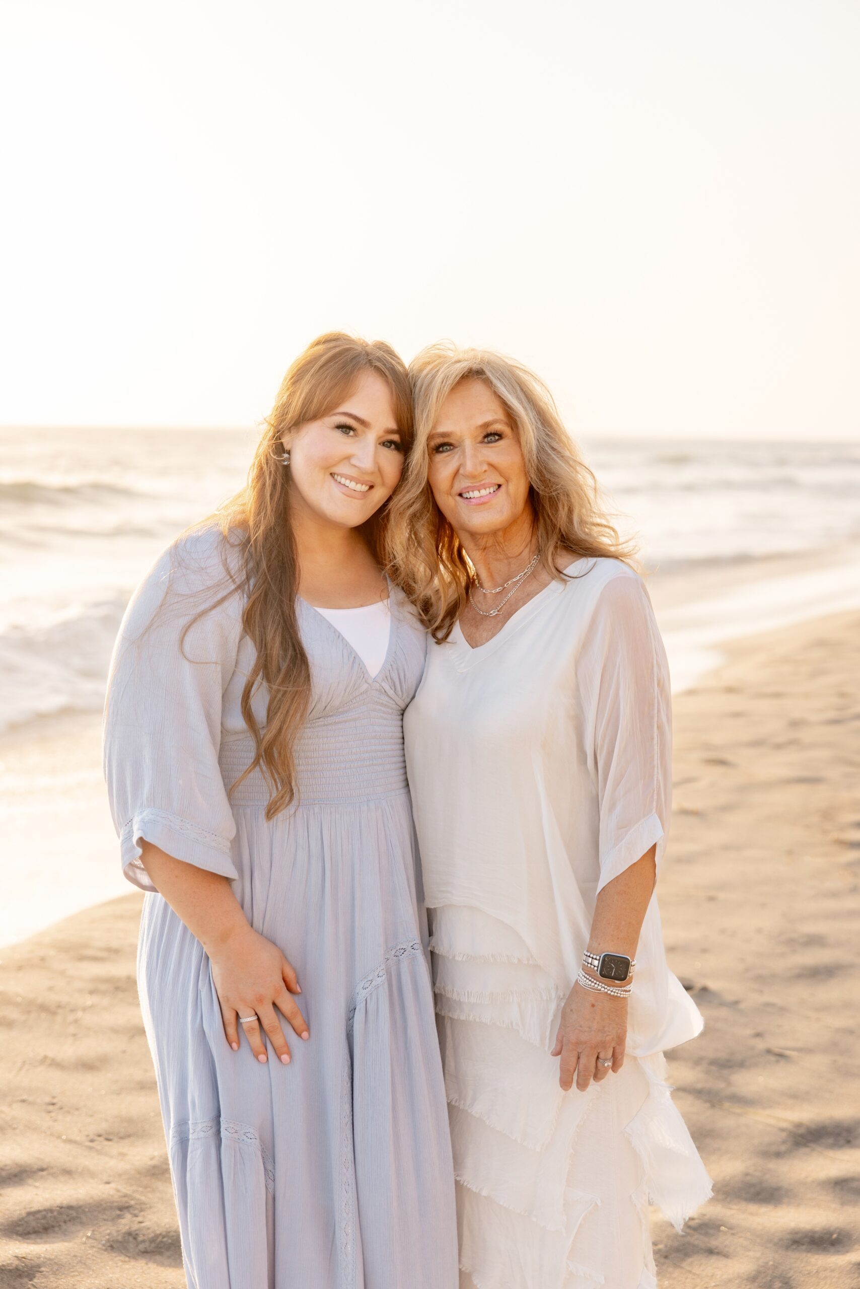 A woman in a blue dress snuggles with mom in a white dress on a beach at sunset